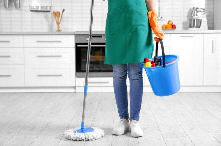 Person in a green apron mopping a kitchen floor with a blue bucket and cleaning supplies, wearing orange gloves.