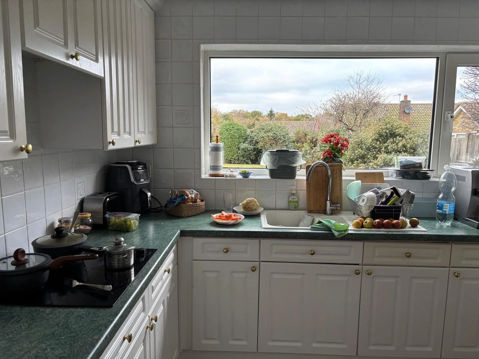 Kitchen with green countertop, white cabinets, window with outside view of trees and houses, various kitchen appliances, dishes, and produce on the counter.