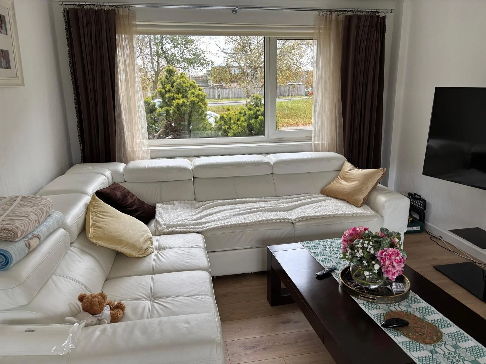 Living room with white sectional sofa, floral centerpiece on a coffee table, large window with trees outside, TV mounted on the wall.