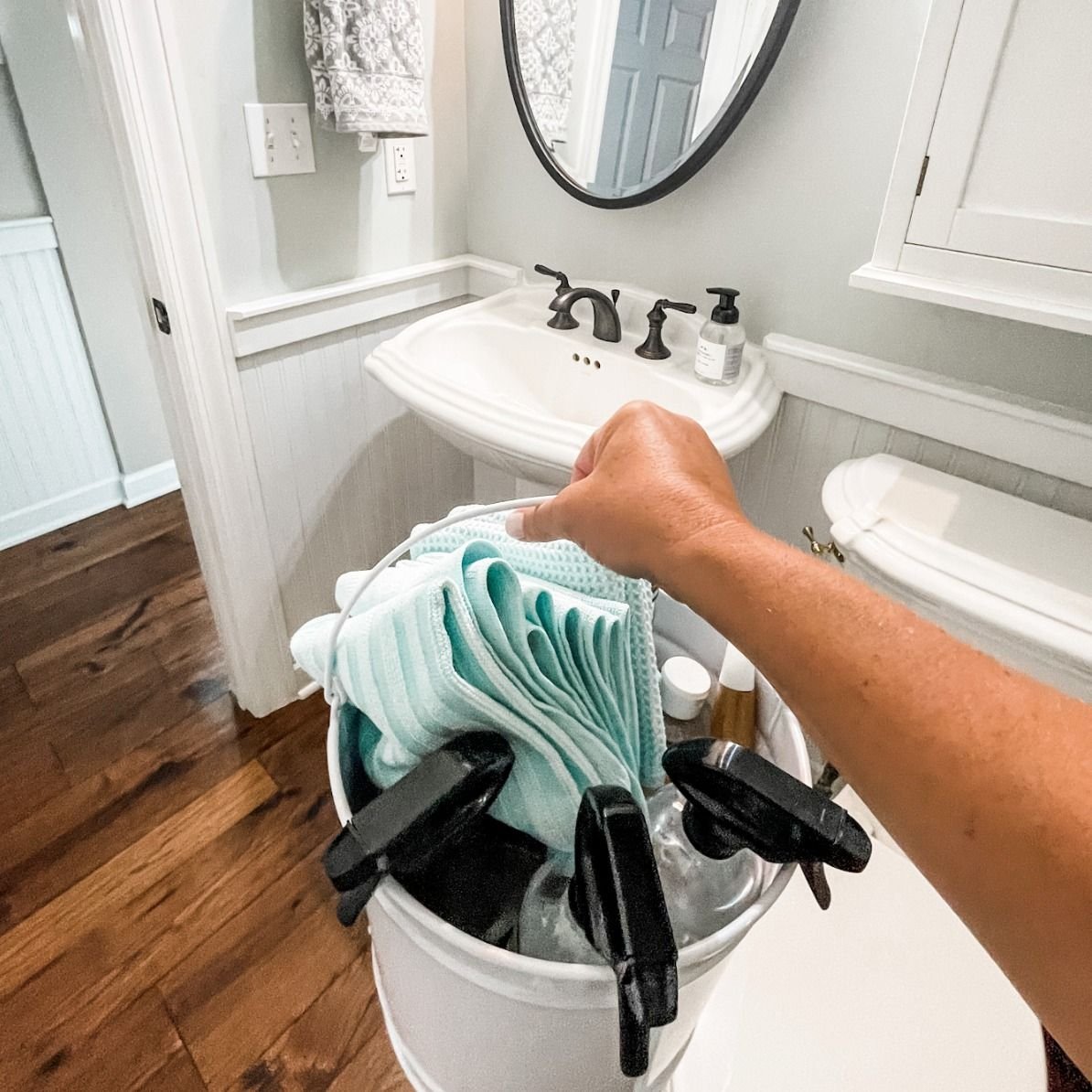 A person reaching into a bucket filled with cleaning supplies, including bleach and cleaning cloths, in a bathroom with a white sink, mirror, and wood flooring.