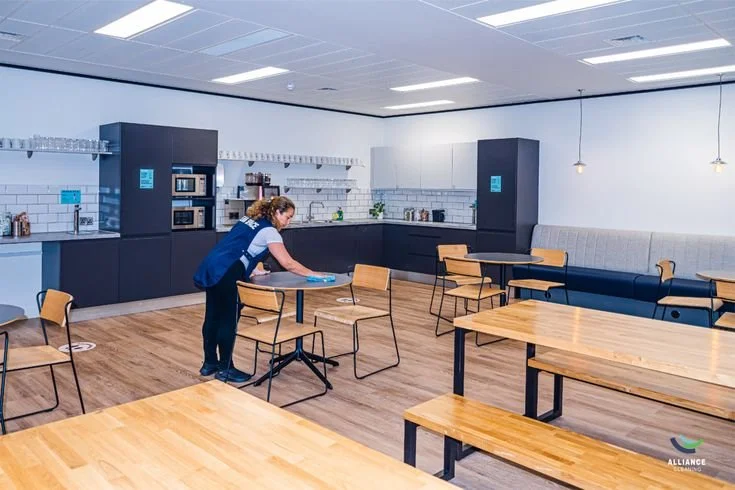 A woman cleaning a table in a modern, brightly lit cafe or lounge area with wooden tables and chairs, black and white kitchen cabinets in the background, and pendant lights hanging from the ceiling.