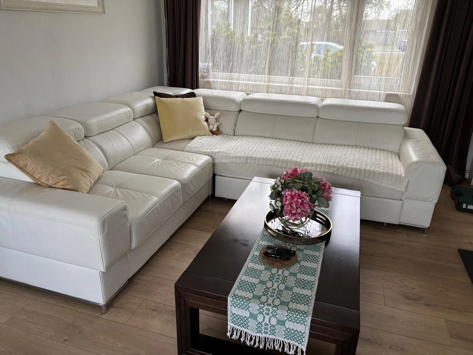 Living room with a white corner sofa, decorative pillows, a wooden coffee table with a floral centerpiece, remote control, and a white blanket, in front of large window with sheer curtains and brown drapes.