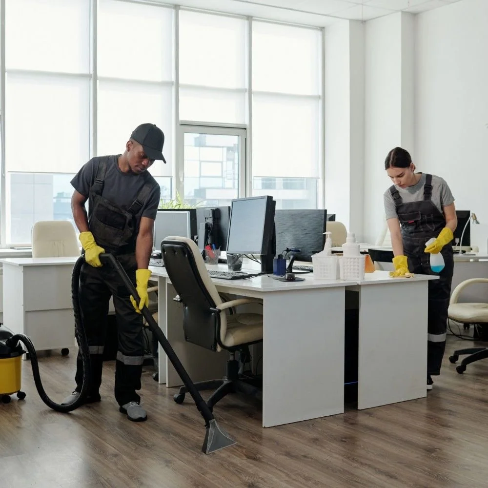 Two cleaning staff, one male and one female, wearing black uniforms and yellow gloves, cleaning an office space with desks, computers, and chairs.
