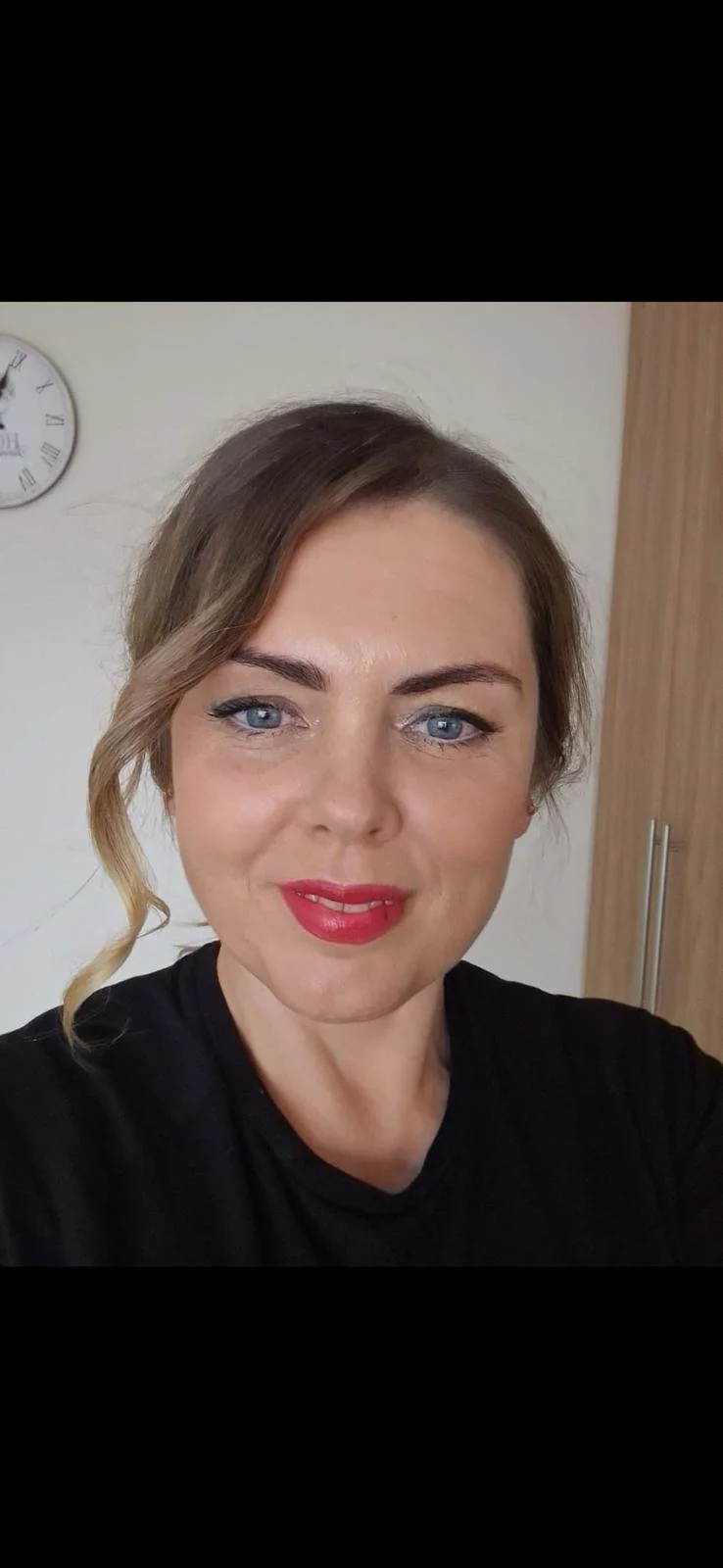 A woman with blue eyes, light brown hair styled to one side with a loose curl, wearing red lipstick and a black top, taking a selfie indoors with a clock on the wall behind her.