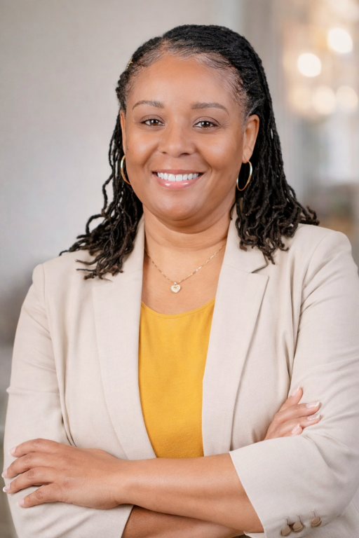 A professional woman with dreadlocks wearing a beige blazer, yellow shirt, and jewelry, smiling with crossed arms in an office setting.