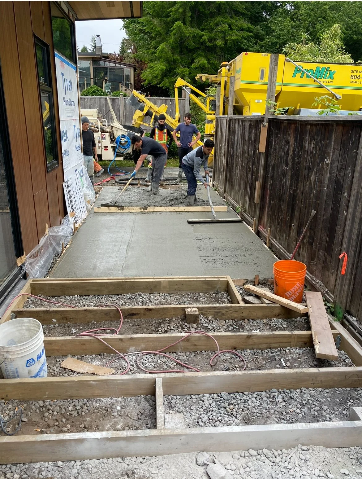 Workers pouring and smoothing concrete on a newly framed patio, with construction equipment and wooden fencing in the background.