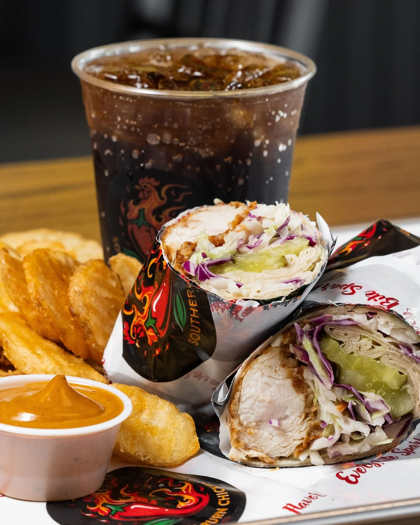 Half of a chicken wrap with shredded lettuce and cucumbers beside crispy potato wedges in a small bowl of sauce, with a glass of soda in the background.