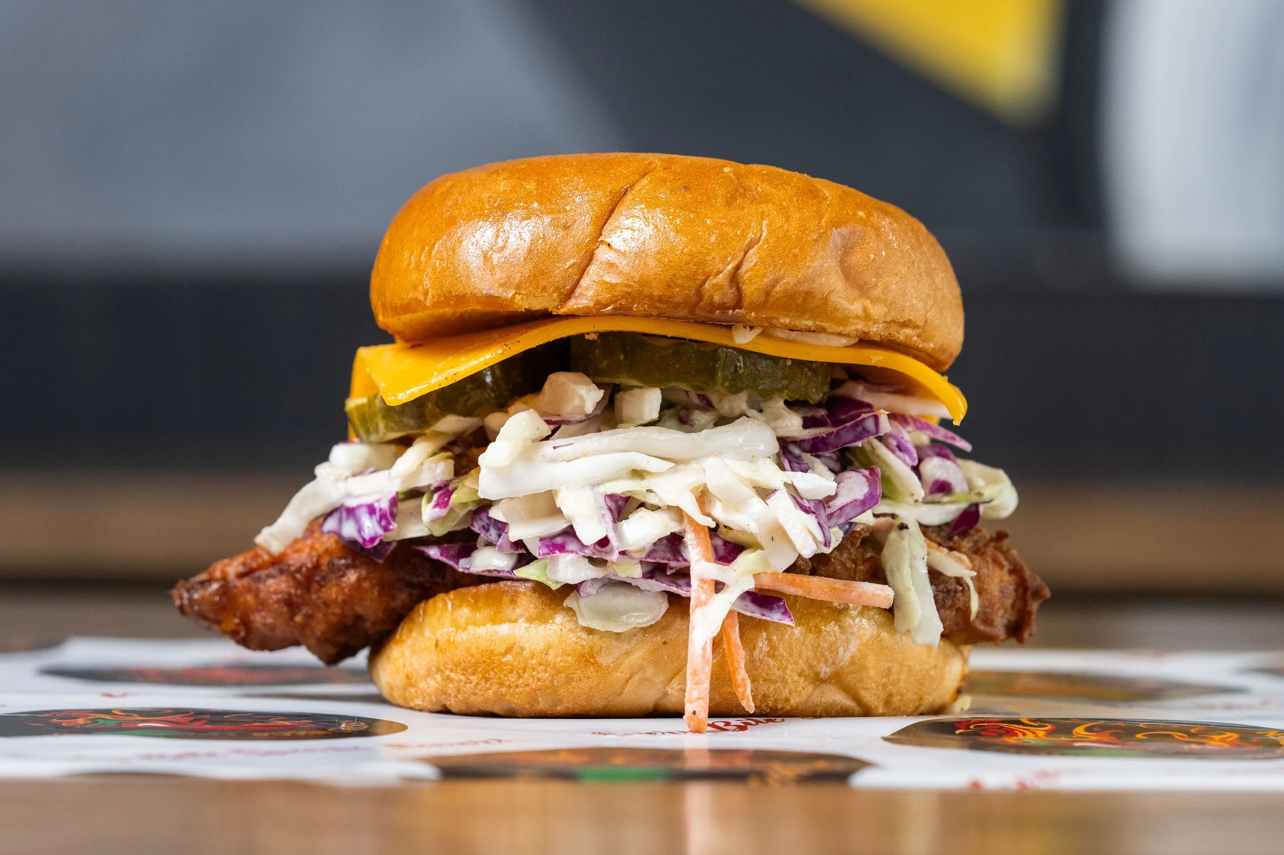 Close-up of a fried chicken sandwich with coleslaw, pickles, cheddar cheese, and a toasted bun on a wooden surface.