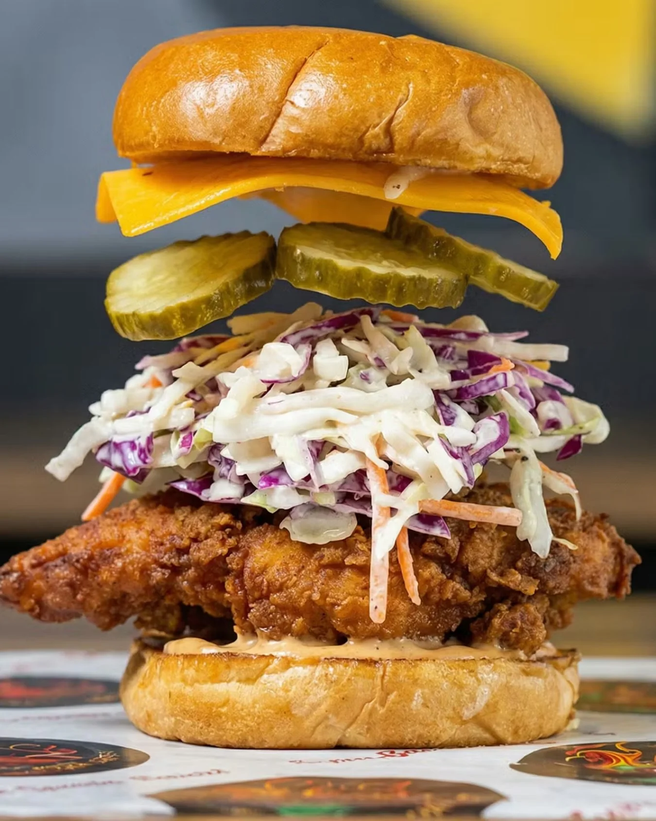 A tall burger with a brioche bun, cheddar cheese, pickles, coleslaw, fried chicken fillet, and bottom bun.