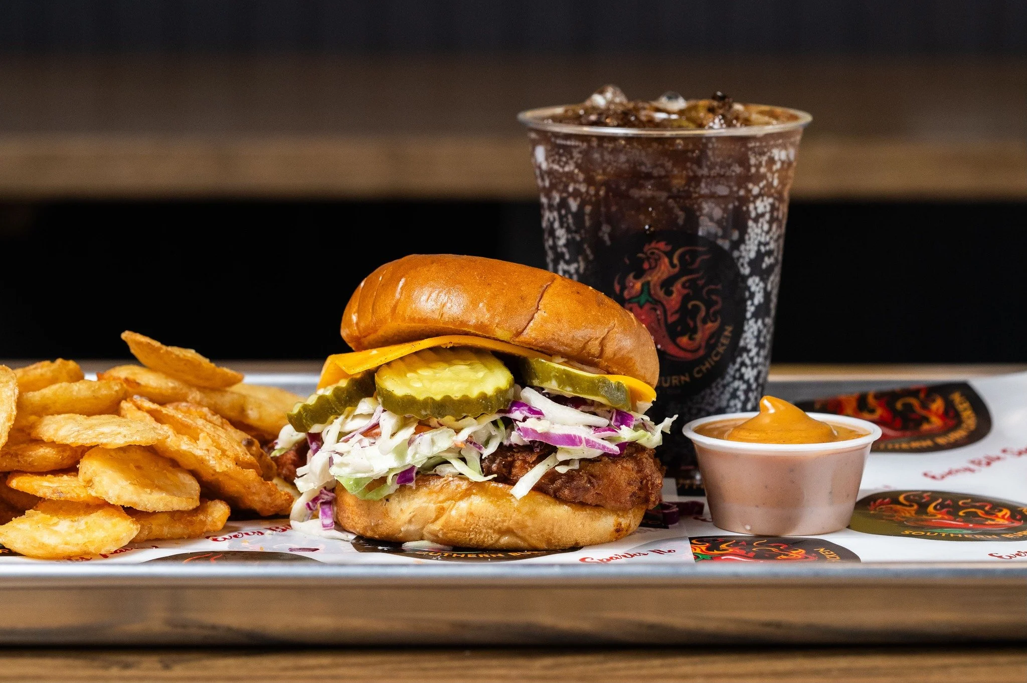 A tray holding a crispy fried chicken sandwich with pickles, lettuce, coleslaw, and a toasted bun, a side of potato chips, a small container of dipping sauce, and a large soft drink in a cup with a black logo.