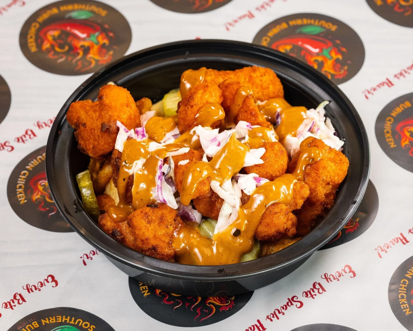 A black container filled with fried chicken pieces topped with shredded lettuce, chopped onions, and drizzled with brown gravy, placed on a branded paper with red and yellow text.