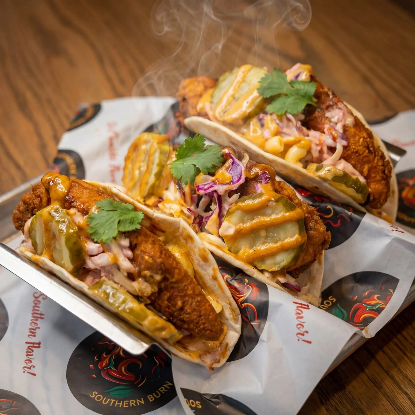 Three steaming hot fried chicken tacos topped with pickles, shredded purple cabbage, cilantro, and drizzled with sauce, served on branded paper in a metal tray on a wooden table.