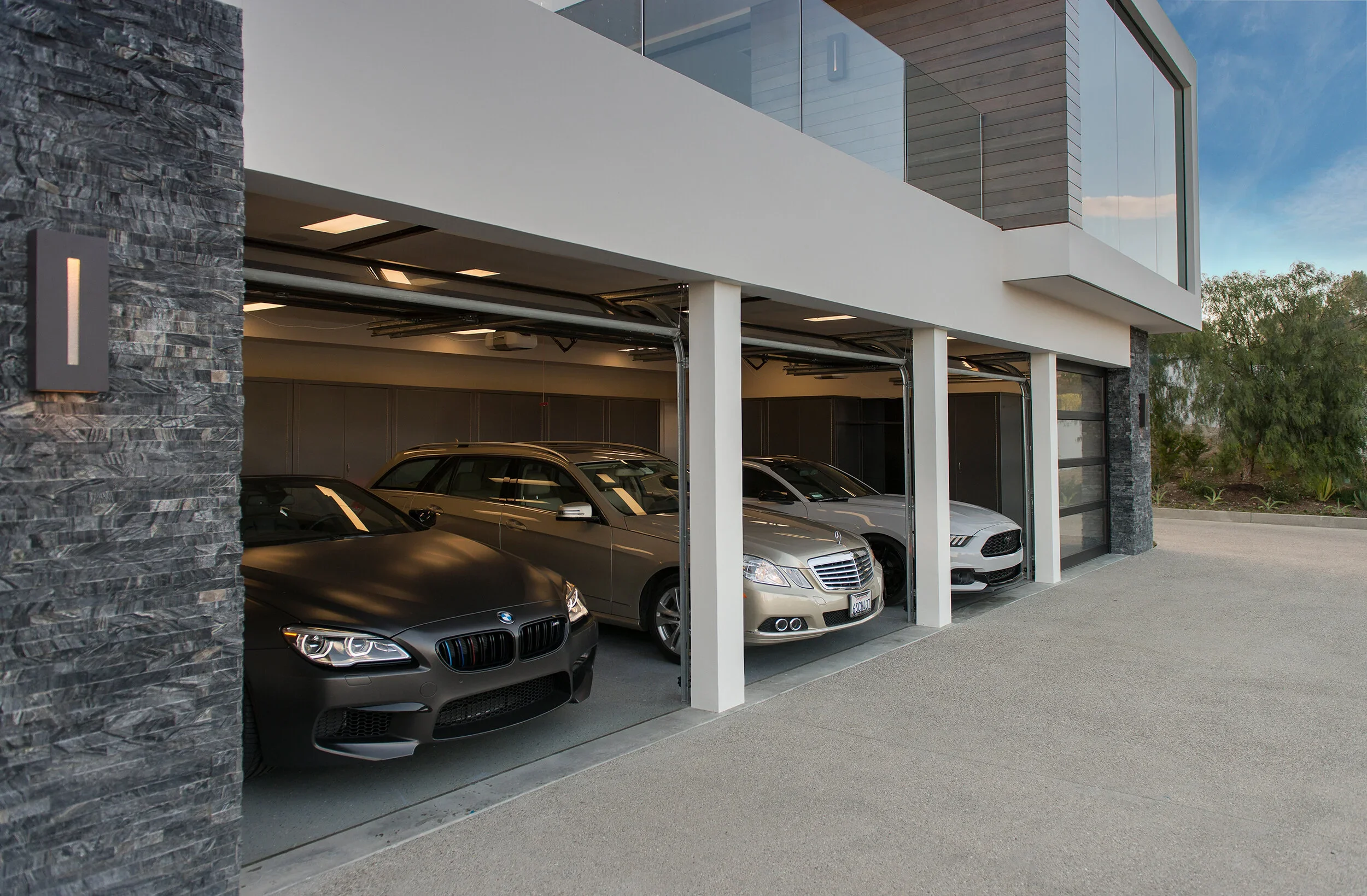 Modern house with a garage containing four parked cars, including a BMW, Mercedes-Benz, Ford, and Mustang, with a mix of stone and white exterior walls and large glass windows.
