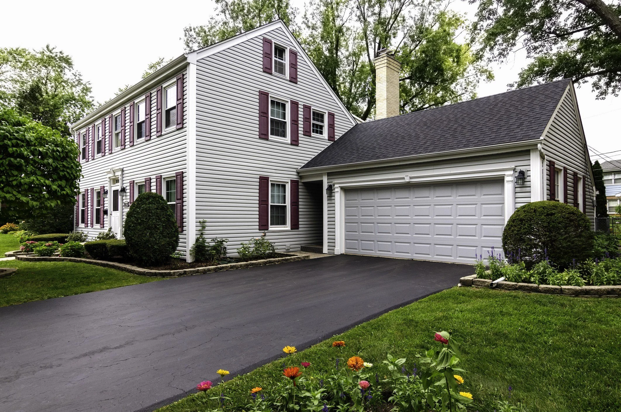 White two-story house with attached garage, black asphalt driveway, and landscaped front yard with flowers and bushes, surrounded by trees.