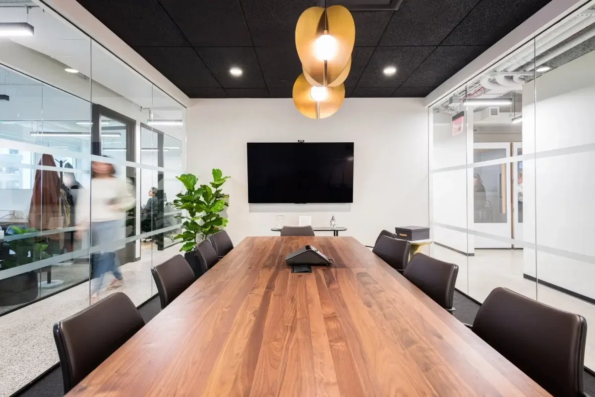 A modern conference room with a large wooden table, black chairs, a flat-screen TV on the wall, a hanging light fixture, and glass walls.