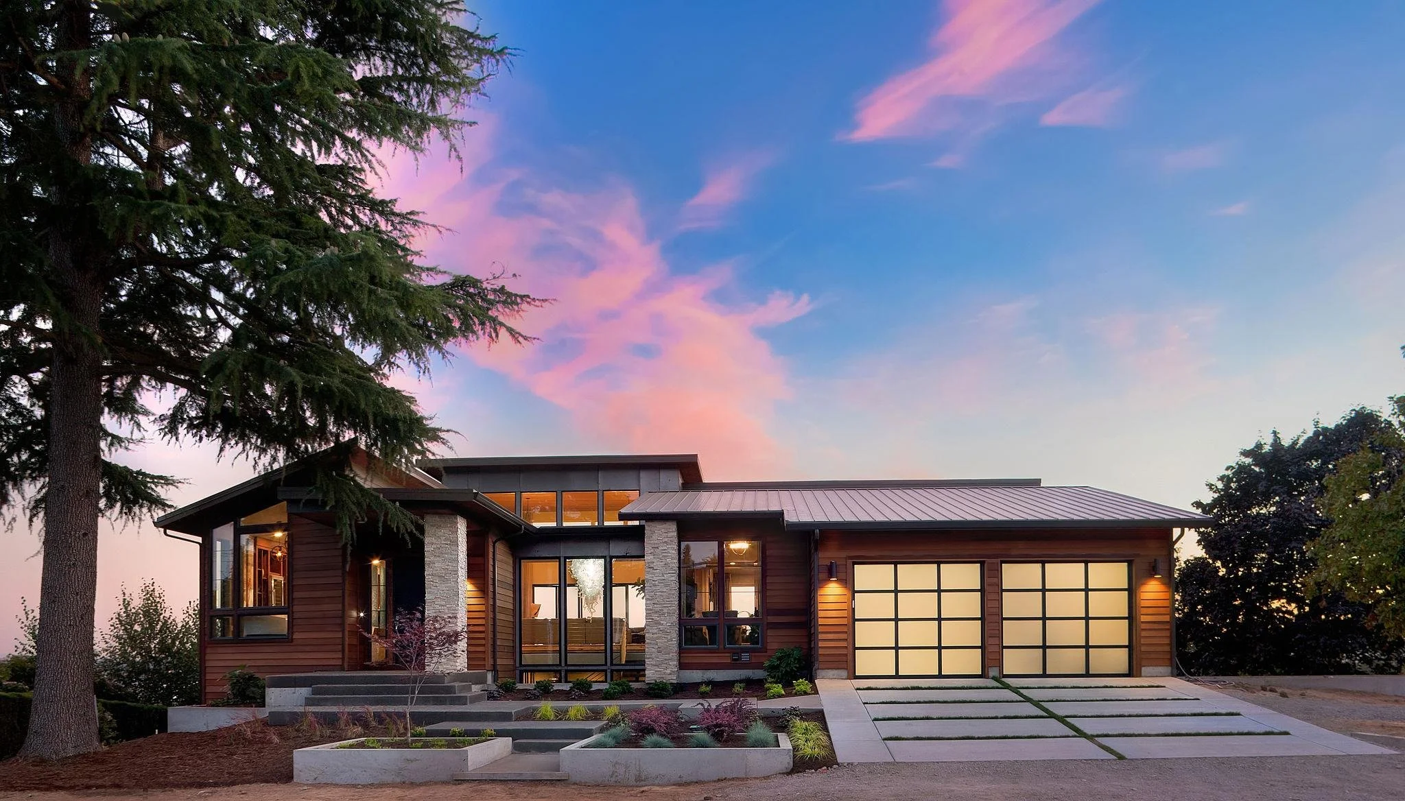 Modern house with large windows and stone accents, surrounded by landscaping and trees, under a colorful sunset sky.