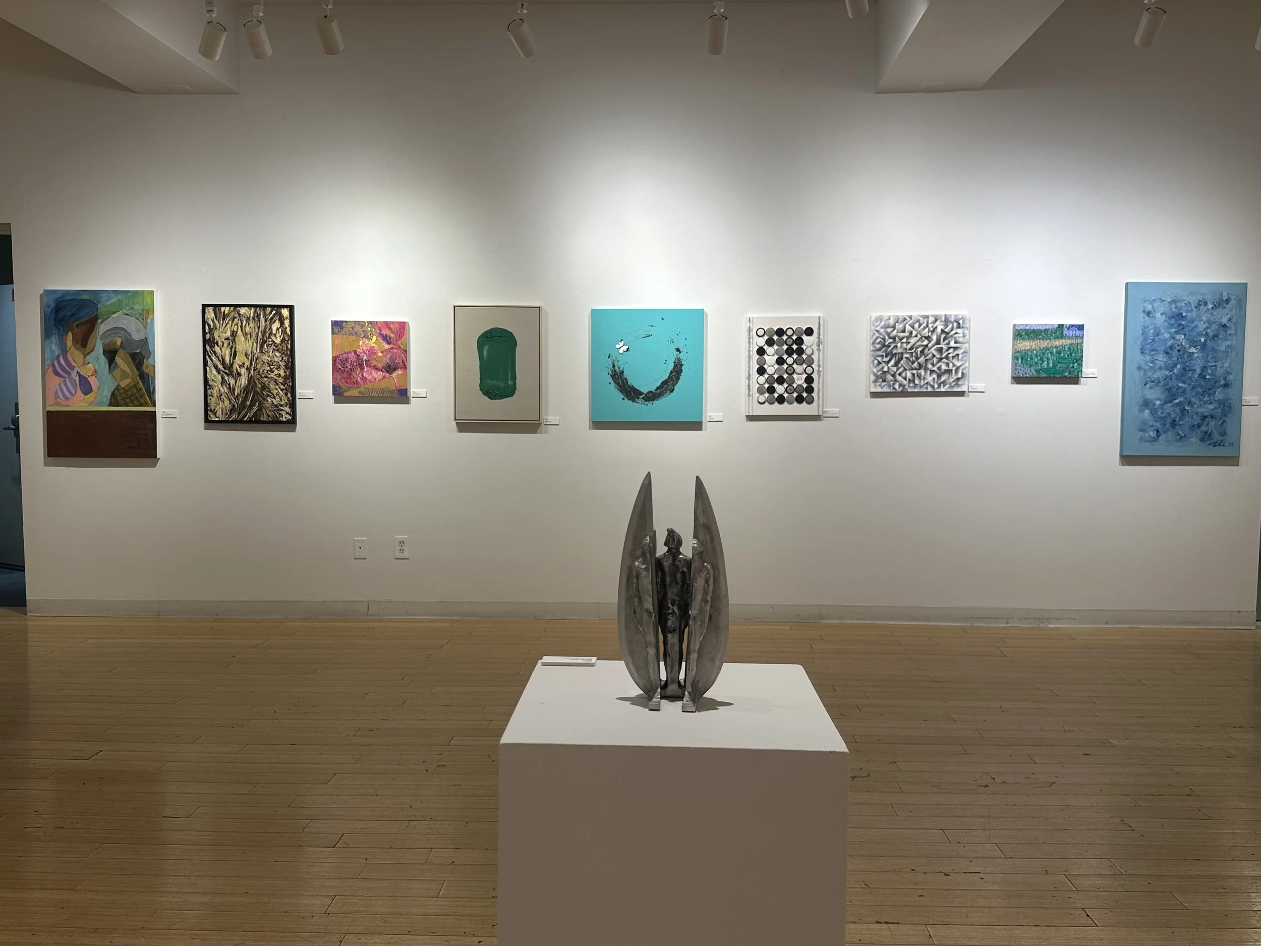 A contemporary art gallery displaying several abstract paintings on the white wall and a small sculpture of a human figure between two large elongated oval-shaped structures on a pedestal.