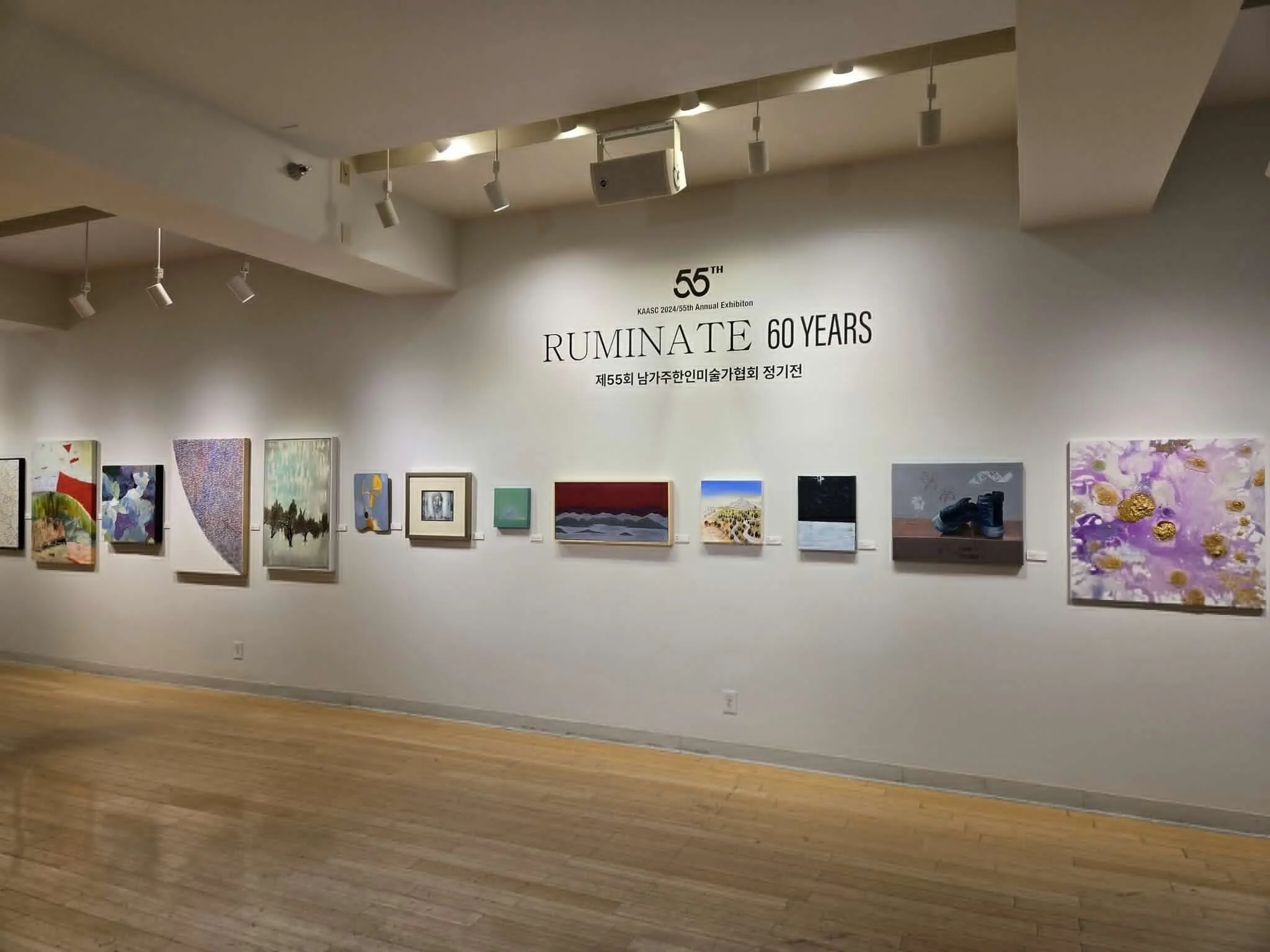An art gallery featuring a row of paintings on a white wall, with gallery lighting overhead. Large text on the wall celebrates the 60th anniversary of the RUMINATE art exhibition.