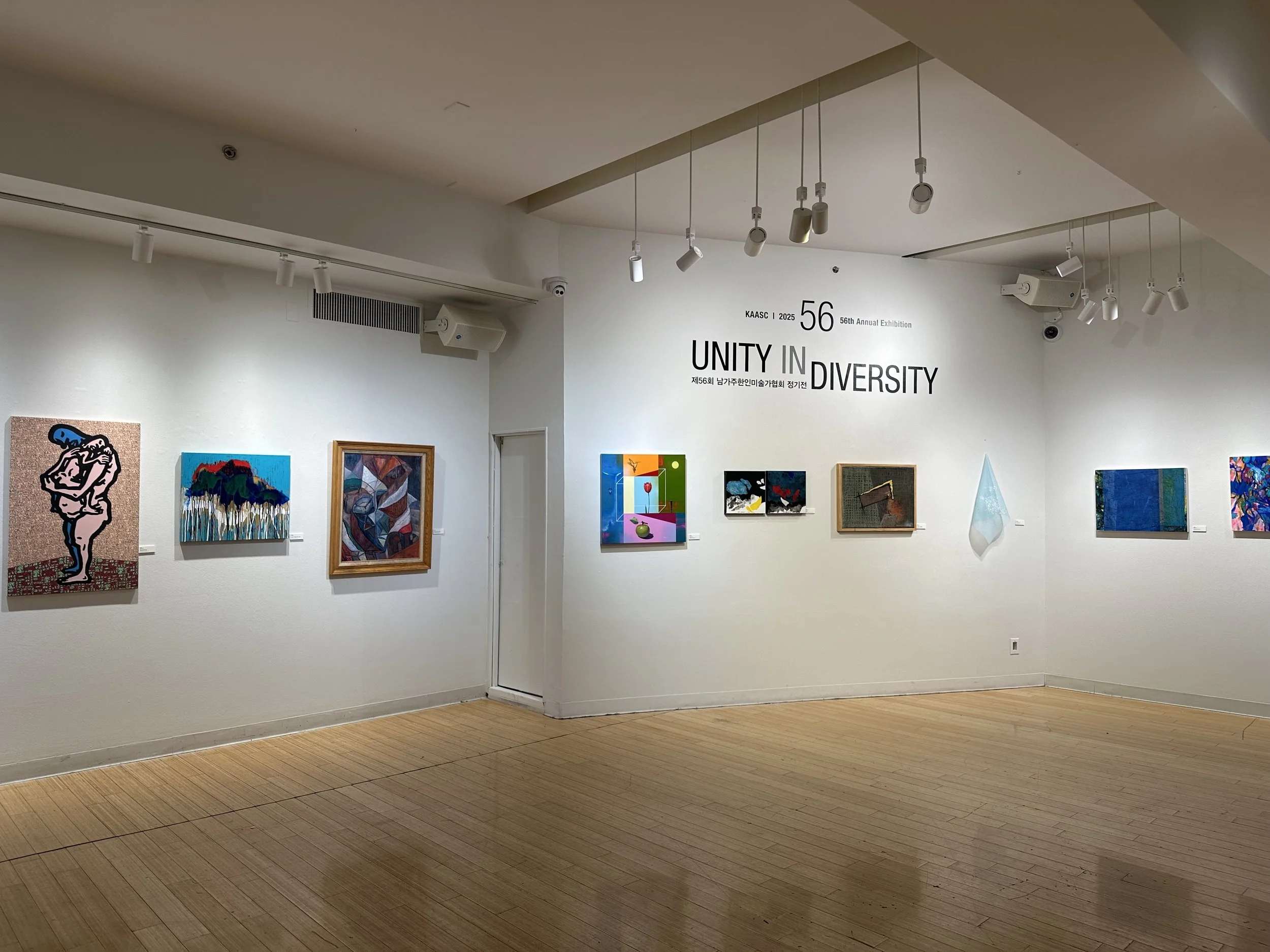 Interior of an art gallery with various paintings on white walls and a wooden floor. There are ceiling-mounted spotlights and a sign that reads "UNITY IN DIVERSITY" along with other text for an exhibition.