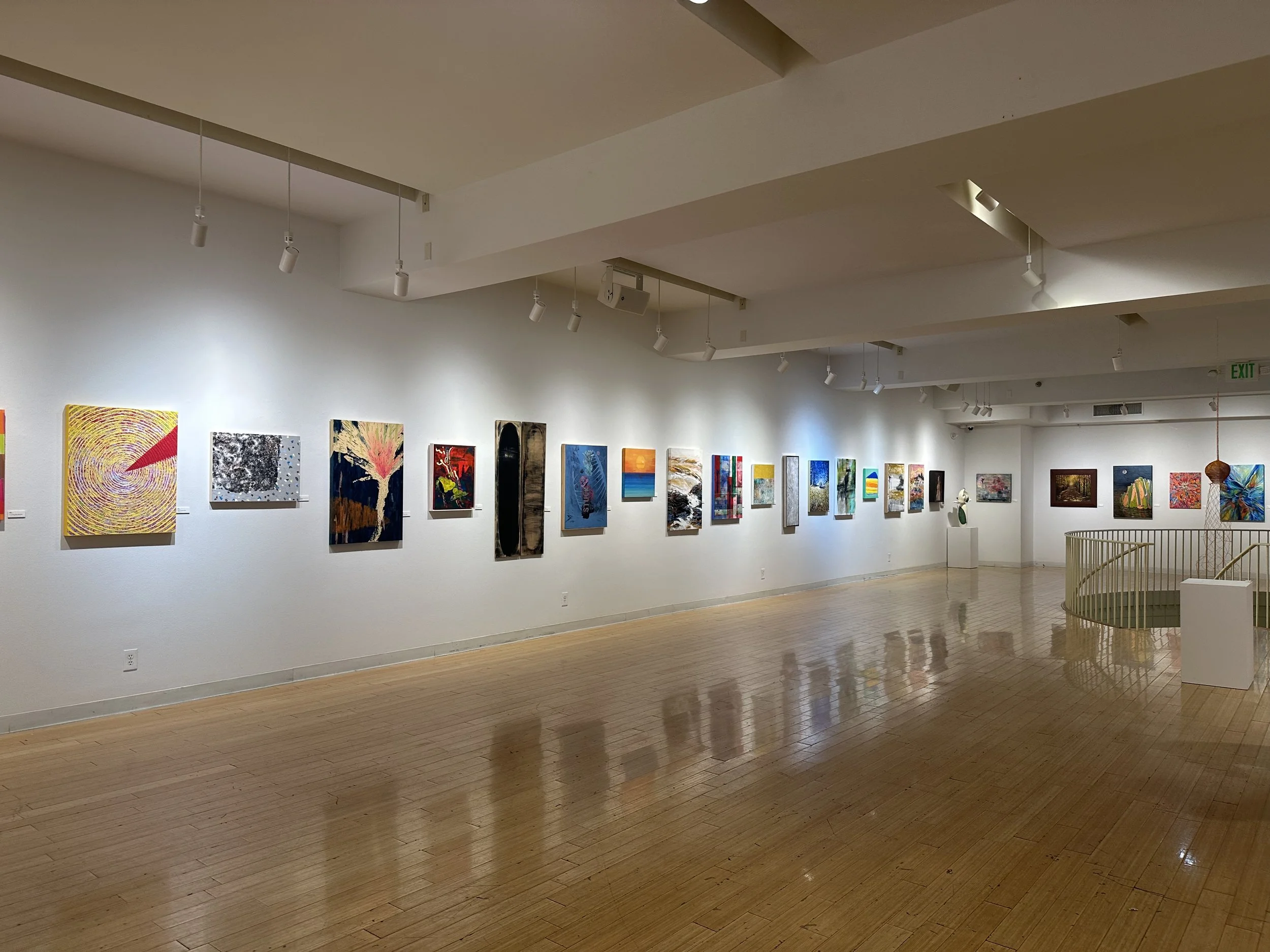 An art gallery with a white wall displaying multiple colorful paintings, hardwood floor, and a spiral staircase to the rightside, with spotlights illuminating the artworks.