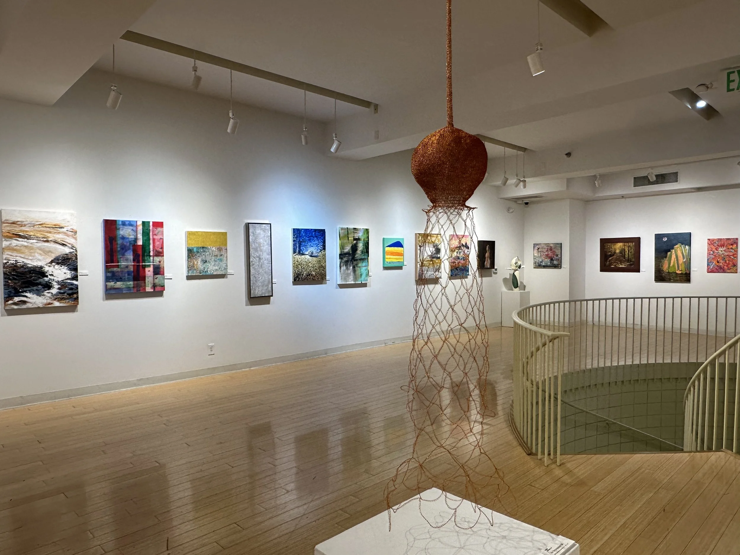 Art gallery with colorful paintings on white walls, a wooden sculpture hanging from the ceiling, and a spiral staircase.