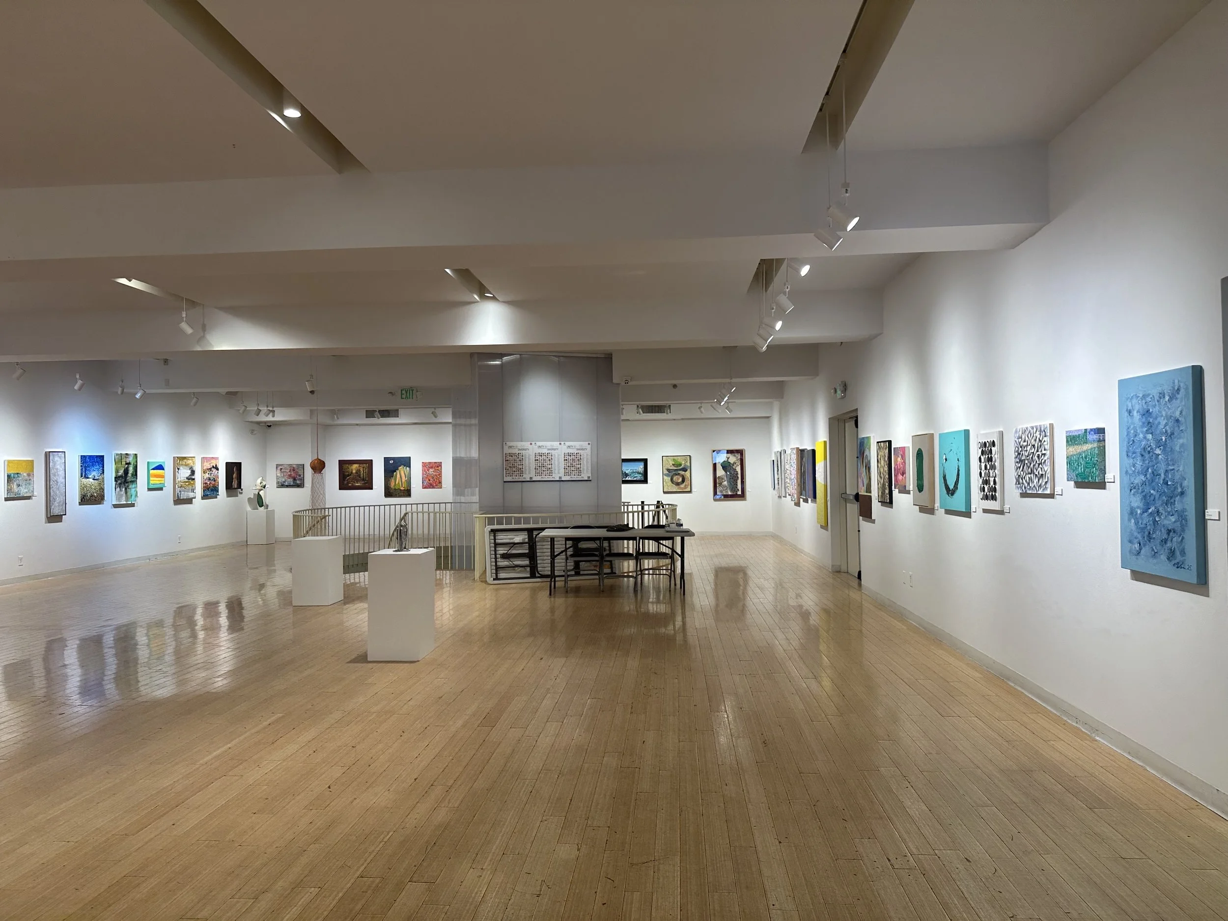 An art gallery with white walls, light wooden floors, and paintings displayed along the walls. There are a few sculptures and framed artworks on the walls, with a central table and some furniture near the back. The space is well-lit with ceiling spot