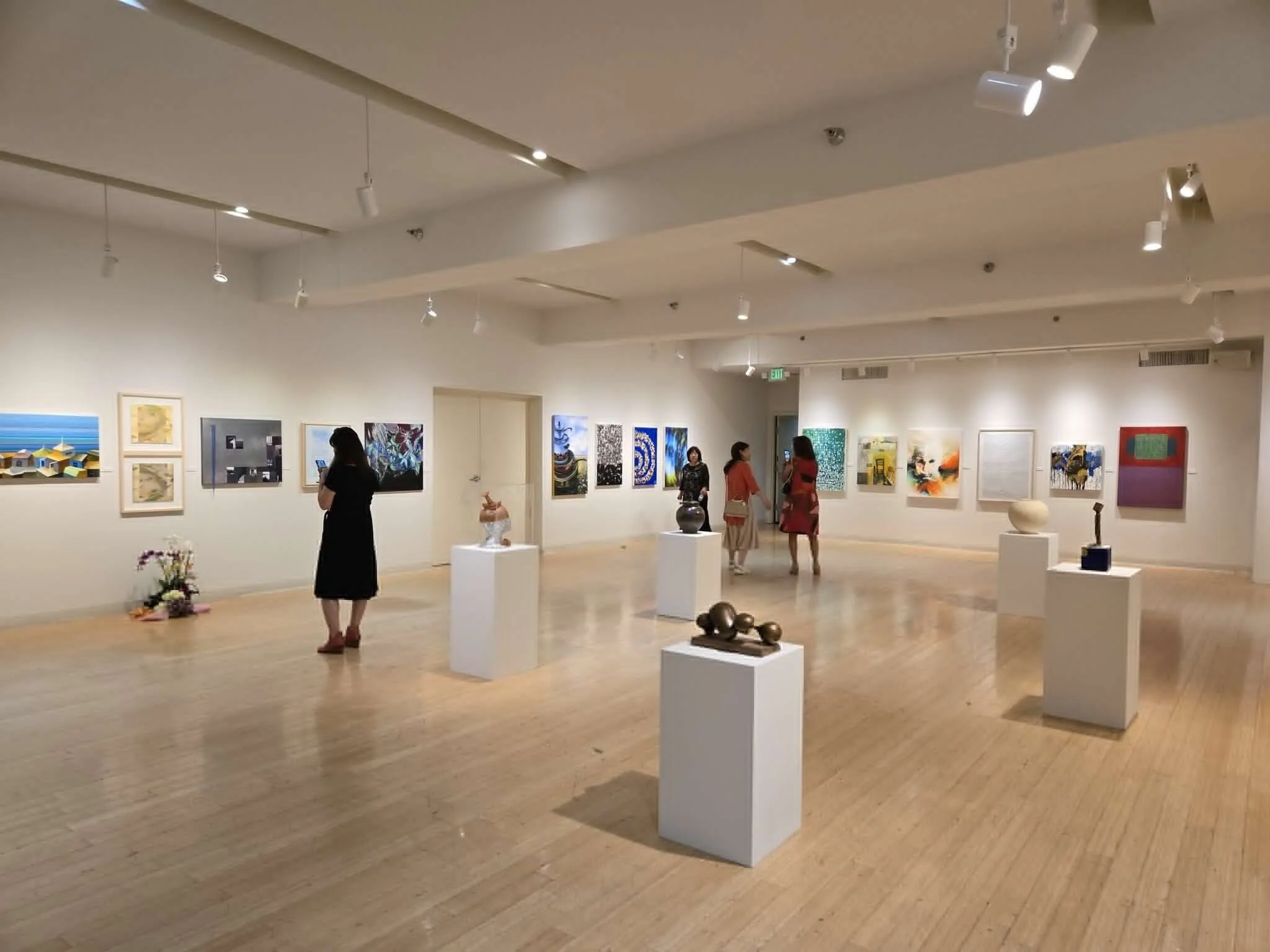 An art gallery with several people viewing paintings and sculptures displayed on white walls and pedestals.
