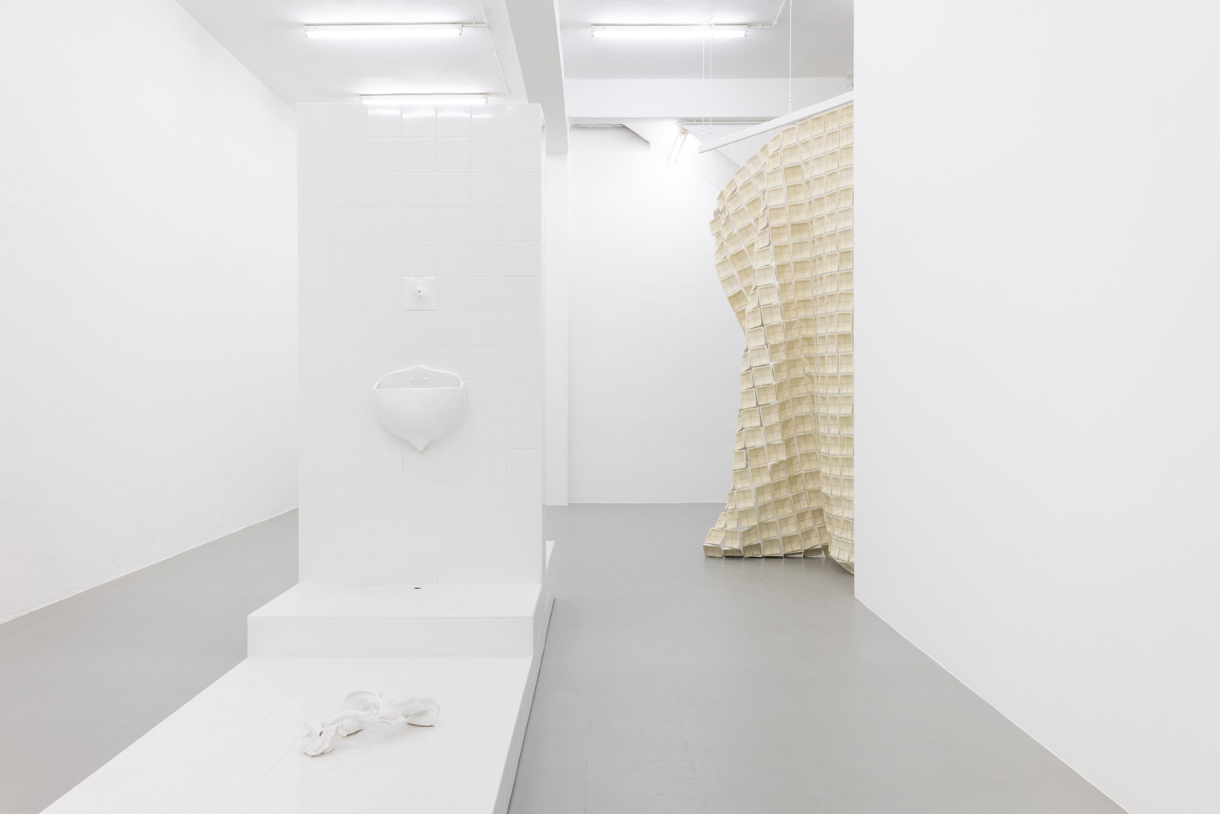 Minimalist art installation featuring a white tiled structure with a urinal and an object resembling a pair of shoes on the floor, with a beige grid-like fabric sculpture to the right in a bright white gallery space.