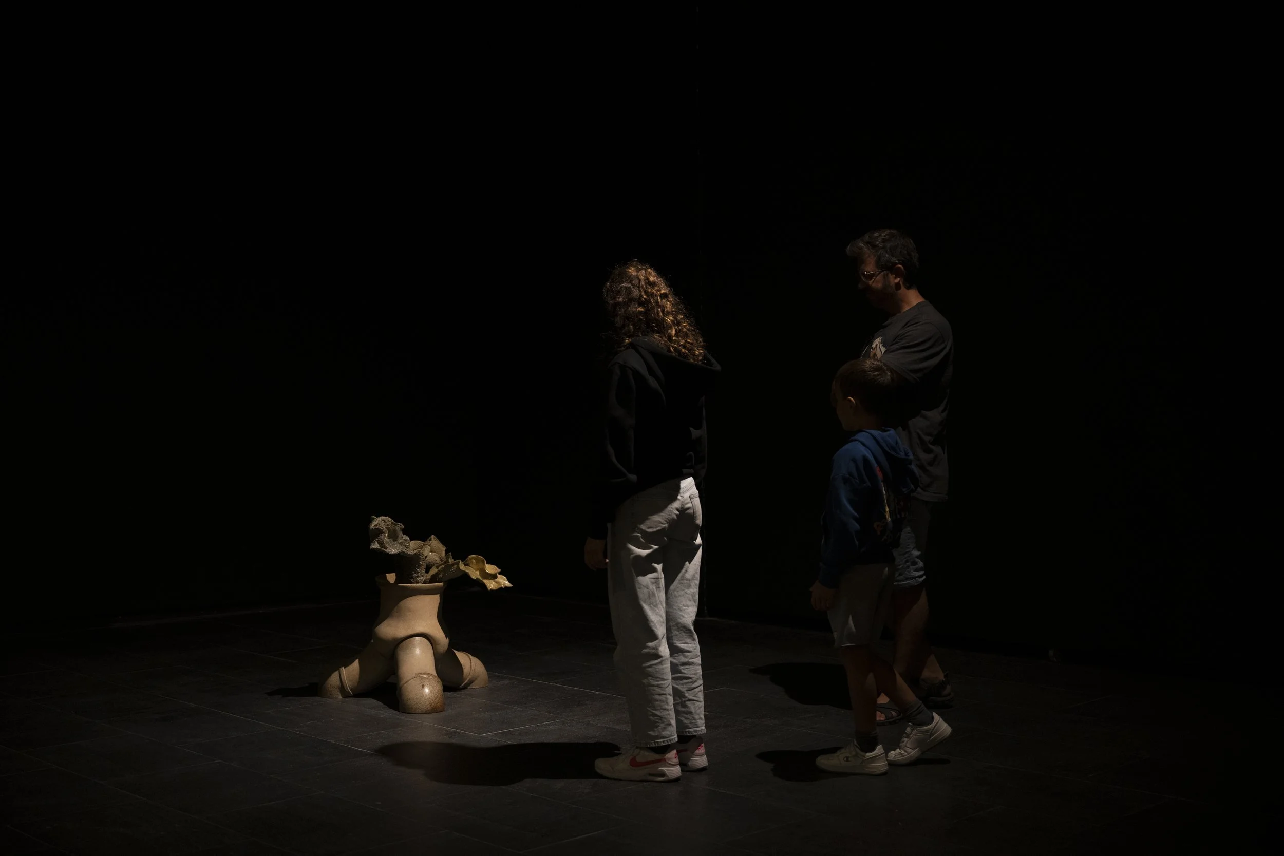 Three people, two children and one adult, standing in a dark room looking at a sculpture. The sculpture resembles a humanoid figure with a wide bowl on its head filled with various objects. The scene is dimly lit, emphasizing the figures and sculptur
