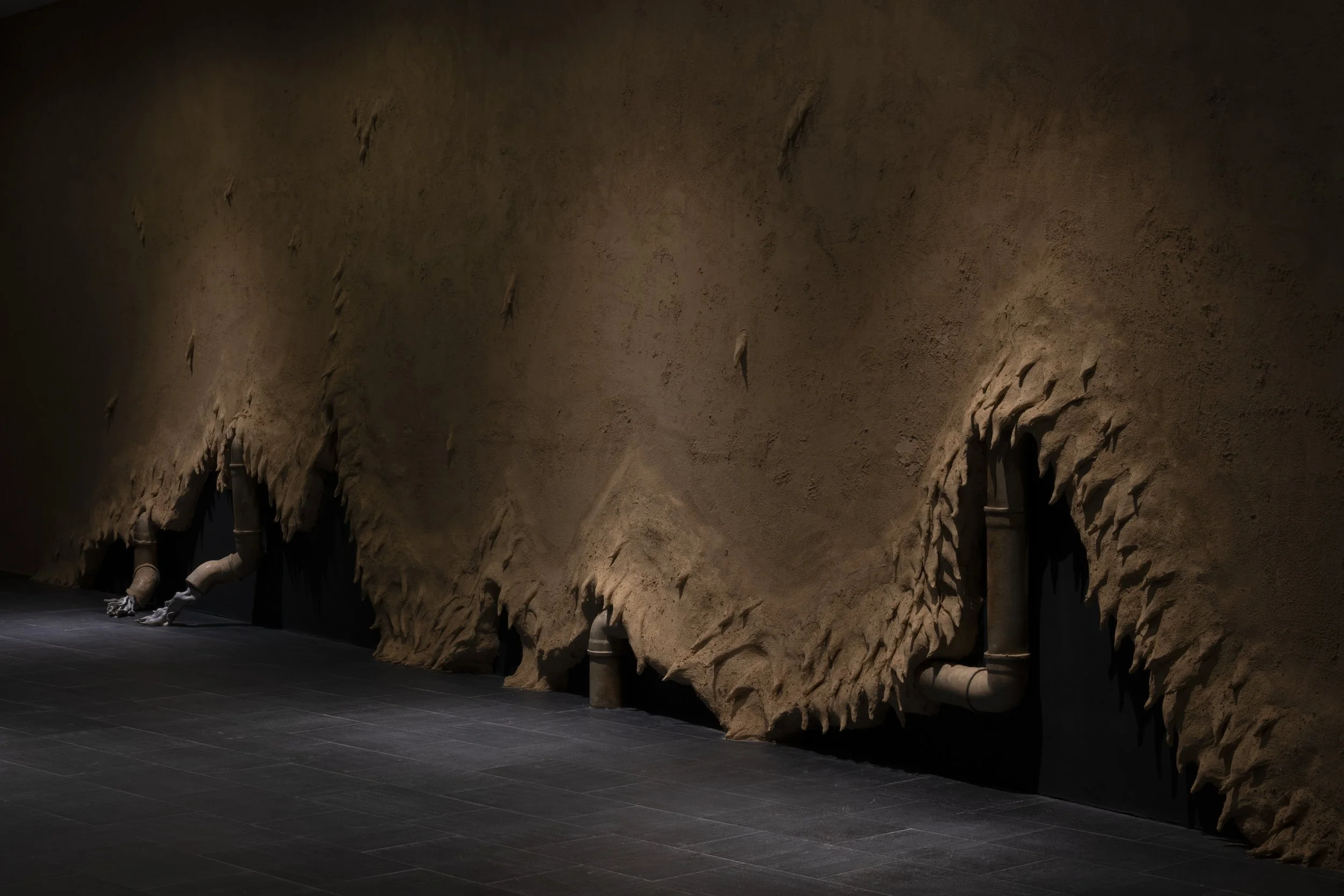 Scene of a room with a textured wall that resembles a cave. The wall has small openings near the floor, connected to pipes. The floor appears to be dark tiled.