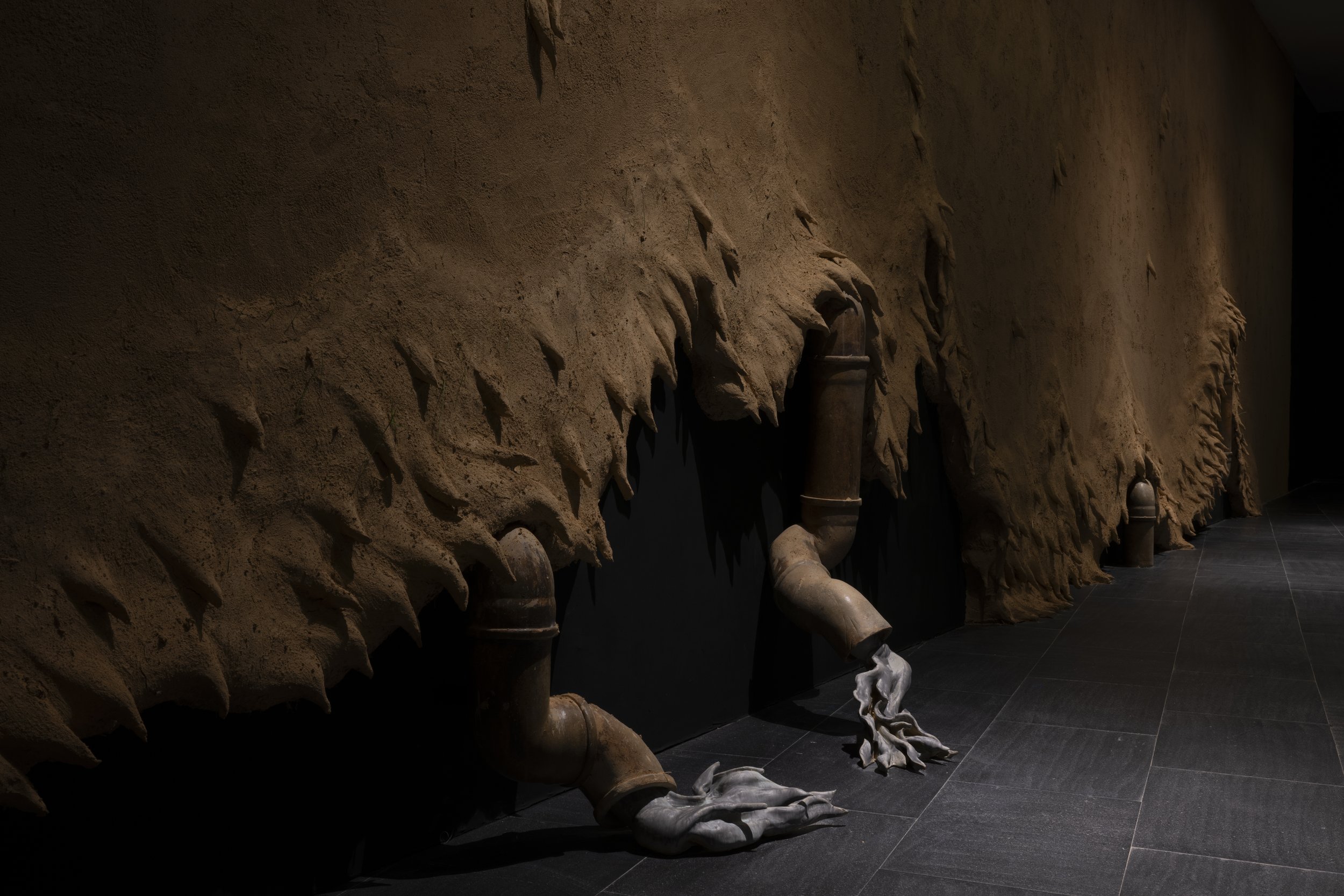 Industrial or decorative installation featuring a large textured beige surface with jagged edges, connected to several rusted pipes with steam escaping from the ends, set on a dark tiled floor.