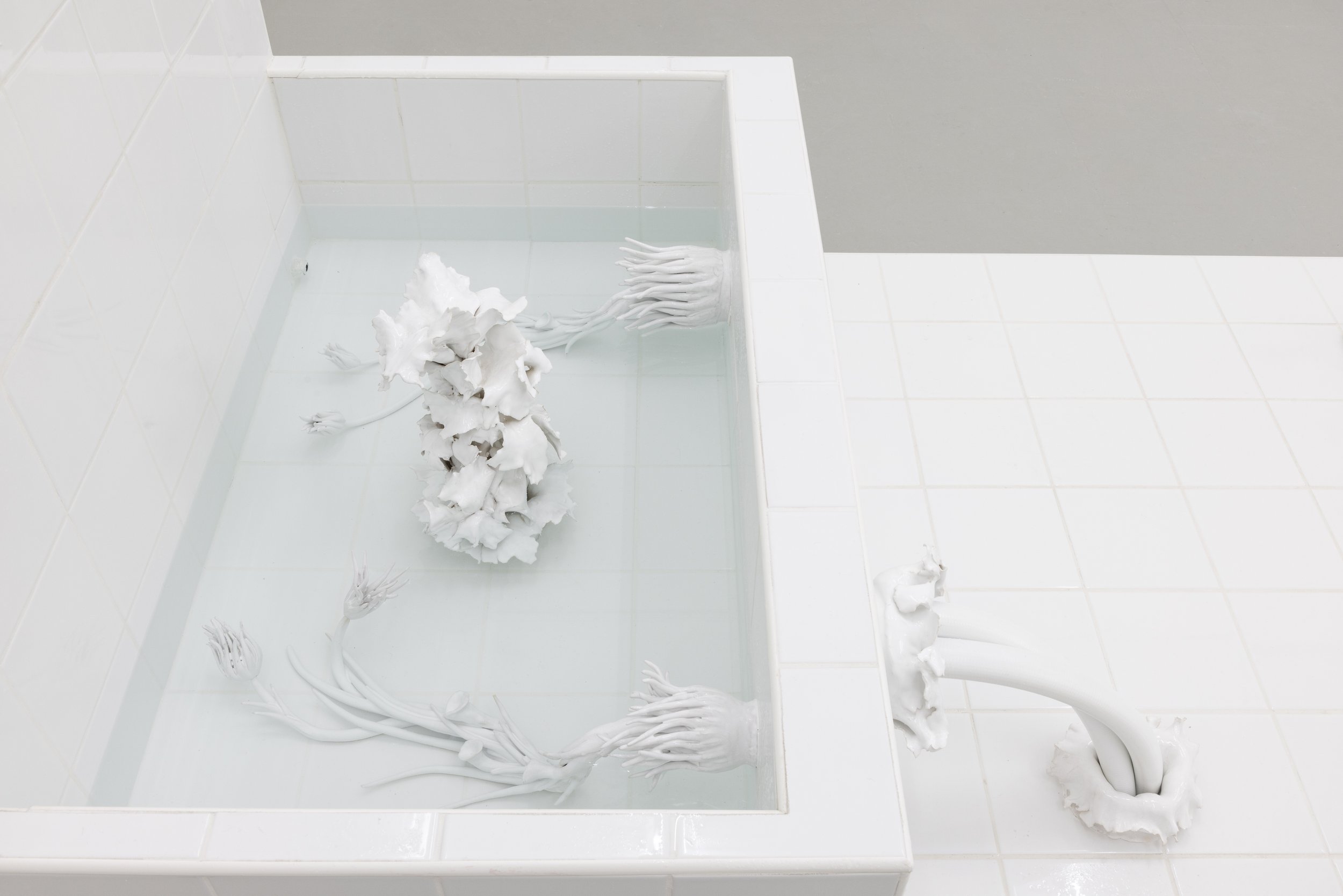 White tiled bathroom with a bathtub filled with water containing white porcelain sculptures of abstract flowers and roots.