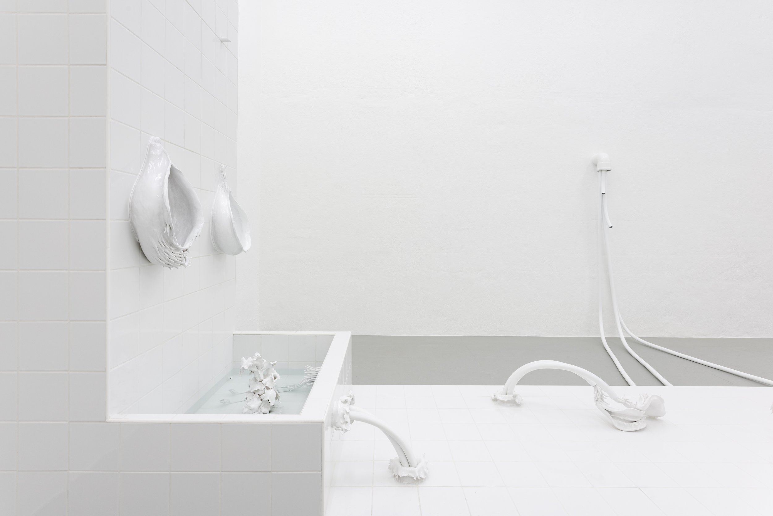 Minimalist white bathroom with tiled wall, hanging washcloths, water fixtures, and a small water basin, all in white.