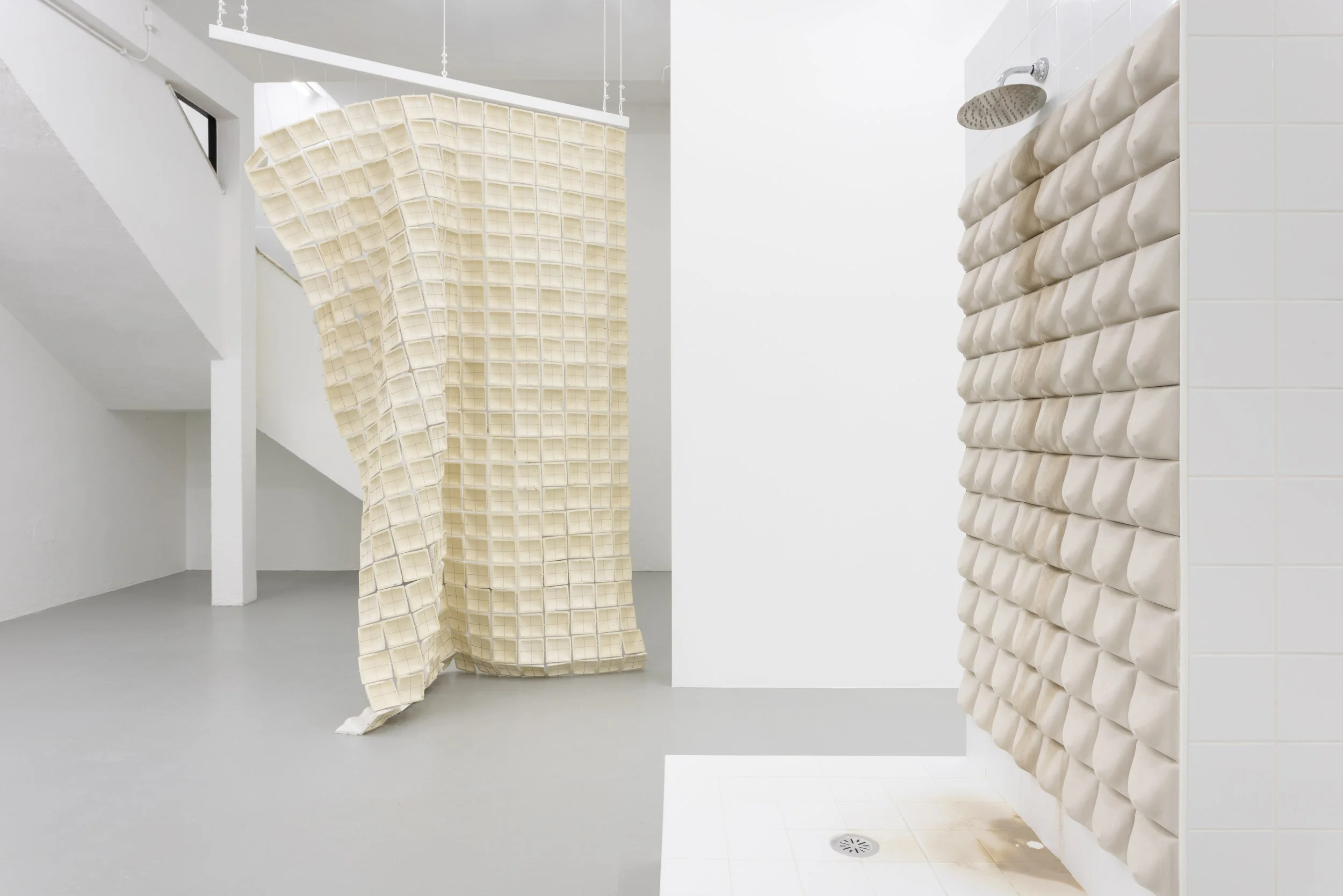 Art installation featuring a hanging sculpture made of numerous small square containers and a wall covered with pillow-like objects in a gallery space.