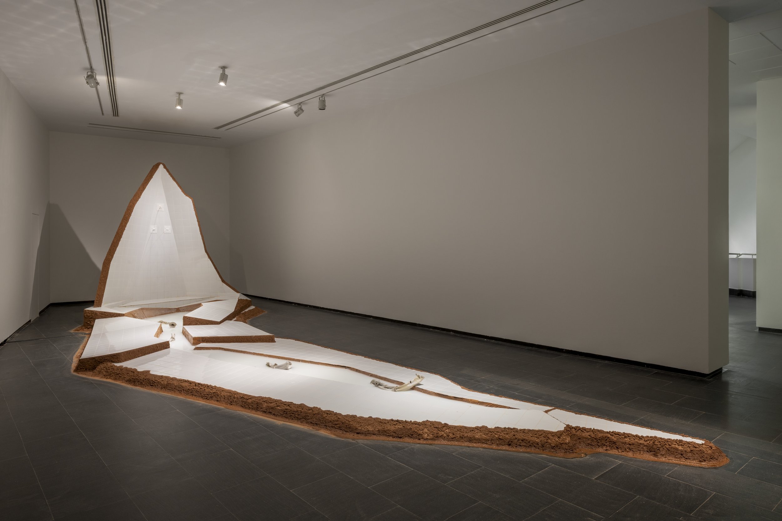 An art installation featuring a white, textured, mountain-like structure with brown edges, laid out on the floor in a gallery space, with a black tiled floor and plain white walls.