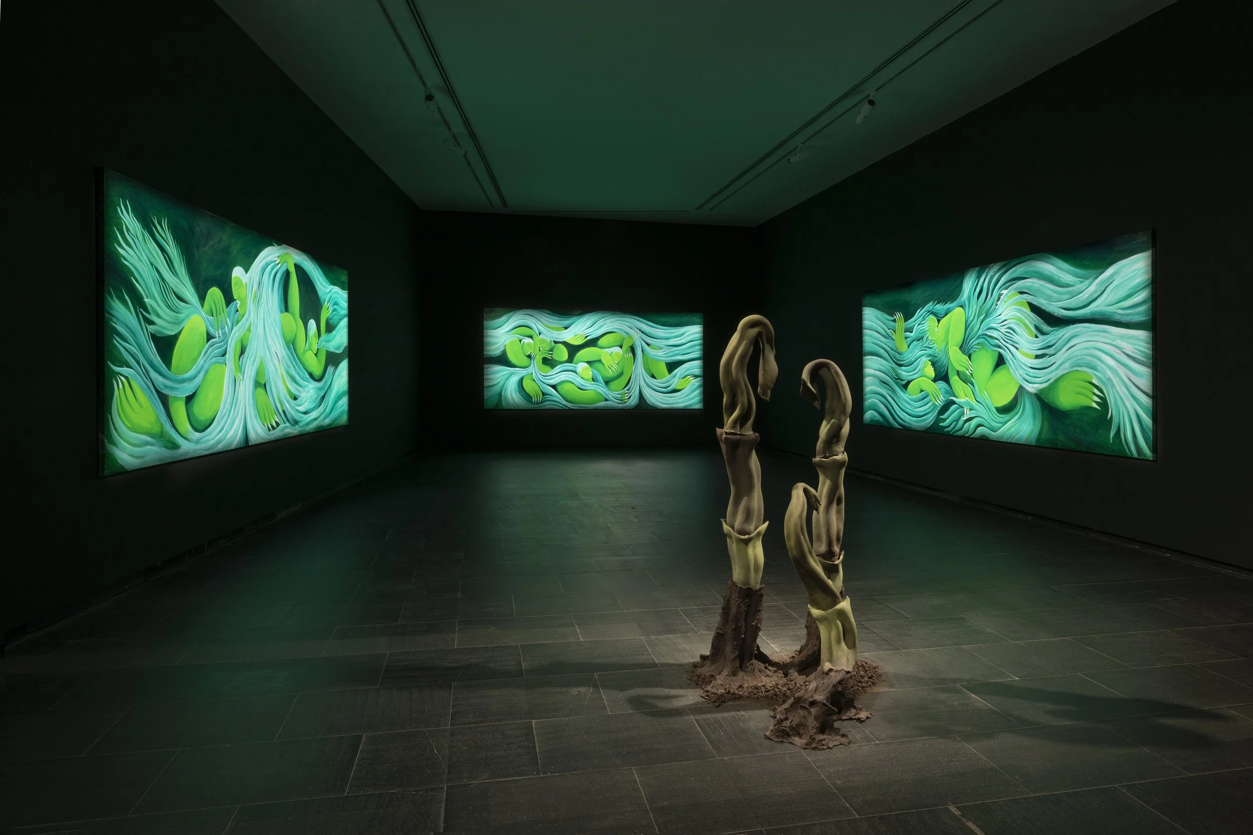 Art gallery with three large digital screen artworks featuring swirling green and blue abstract designs, and a sculptural installation of four elongated, twisted, beige forms on a brown base in the center.