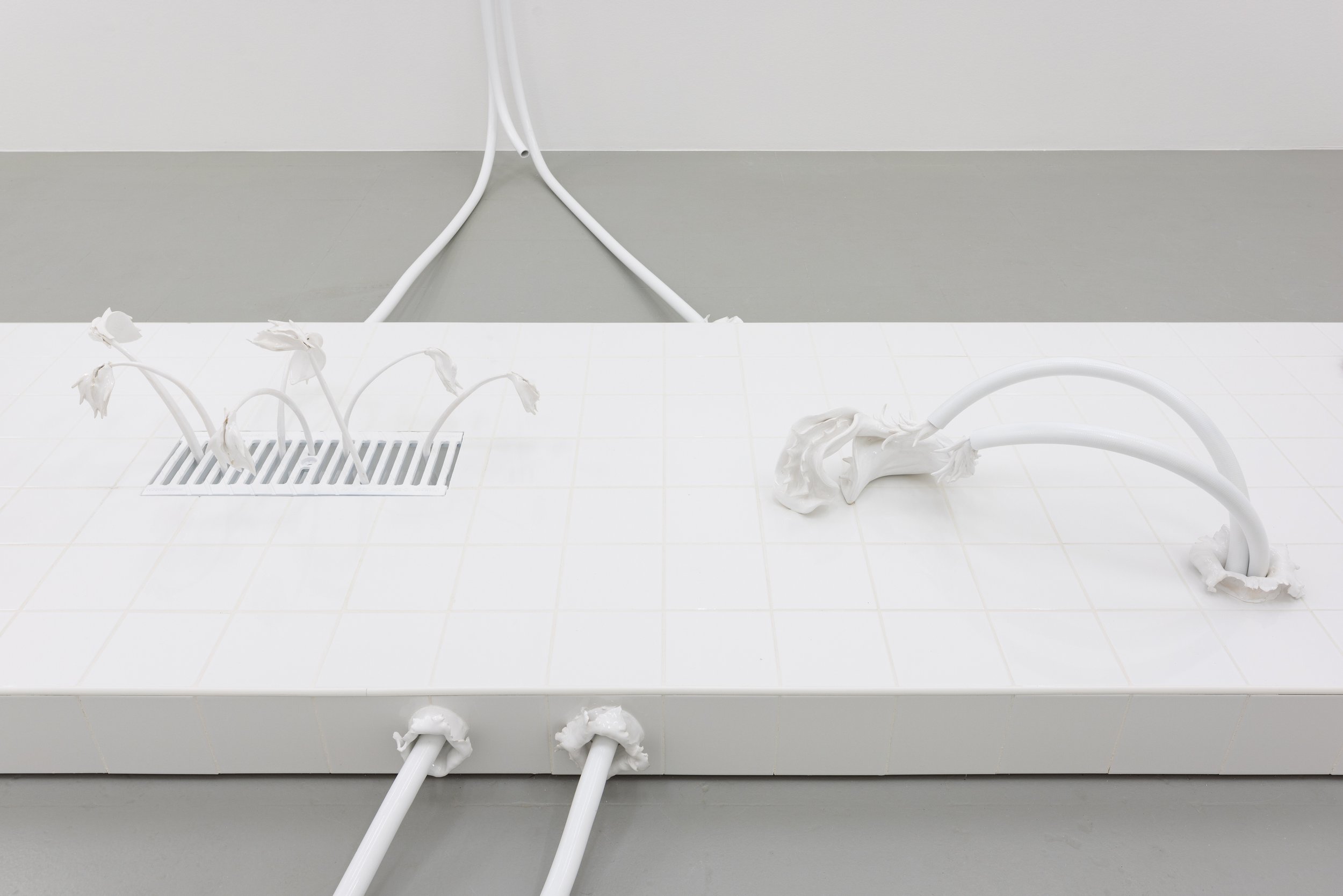 White tiled surface with bent and drooping white fabric tubes resembling flowers, some connected to the wall and others emerging from holes in the tile, with a drain and blinds in the background.