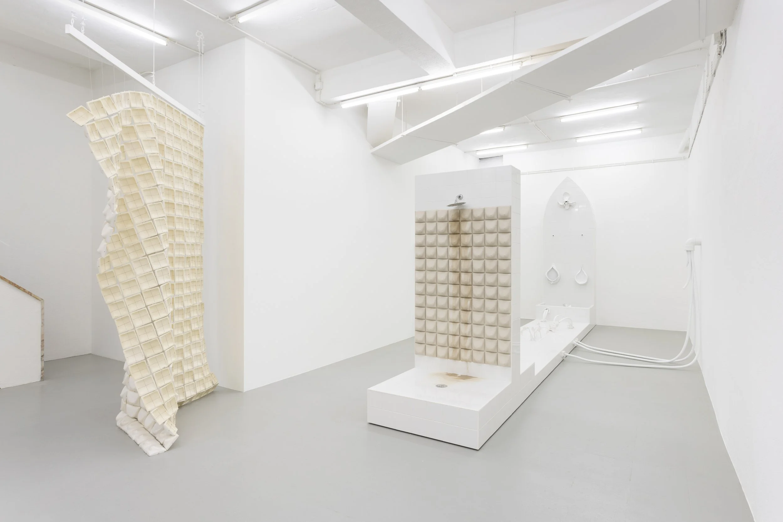 Minimalist art installation in white room featuring a large tower of padded foam cubes, a white platform with textured foam padding, and wall-mounted fixtures with hoses.