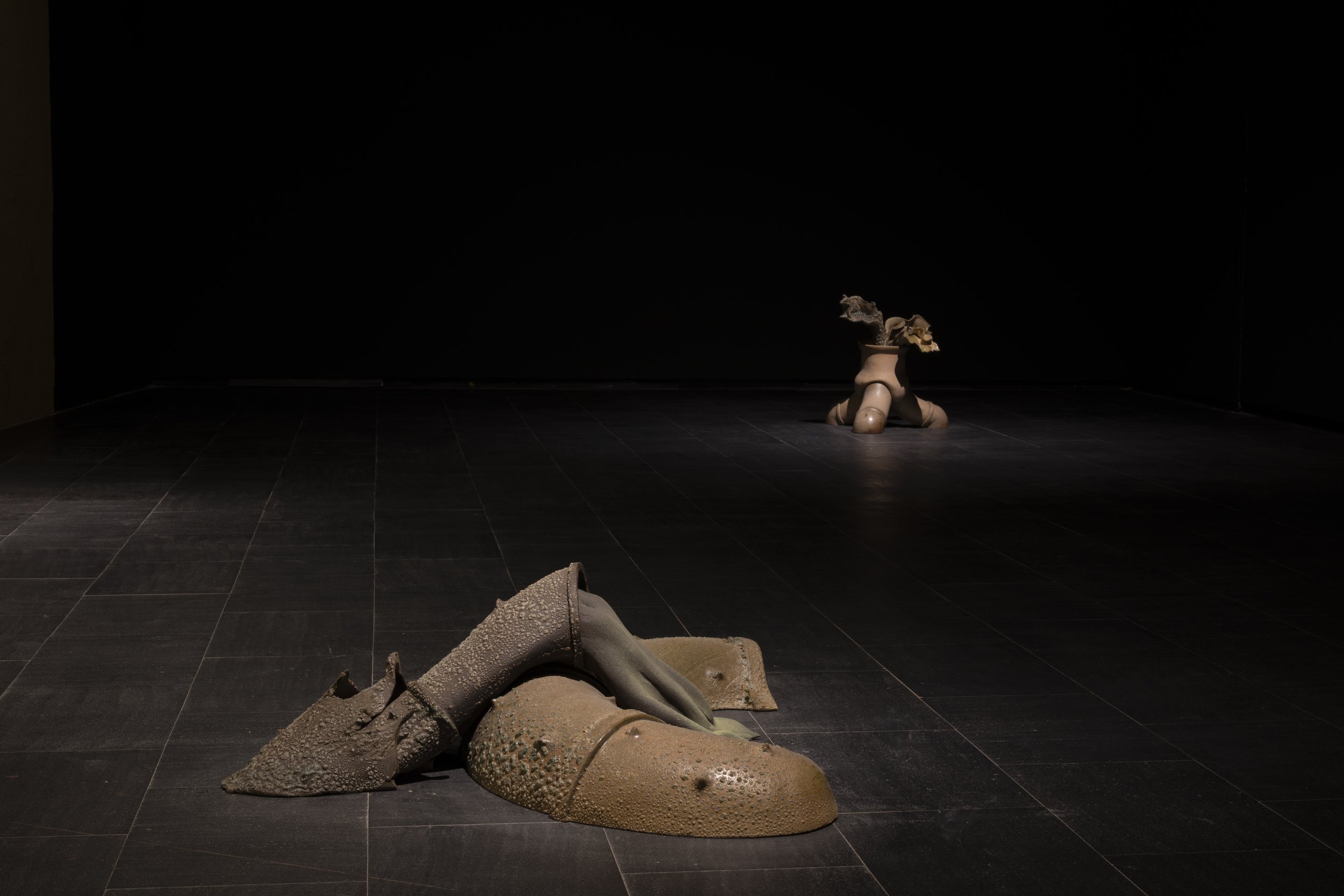 Art installation with two abstract sculptures on a dark floor and black background; one sculpture in the foreground resembles a reclining figure with textured and smooth surfaces, the other in the background resembles a small figure with a cloud-like