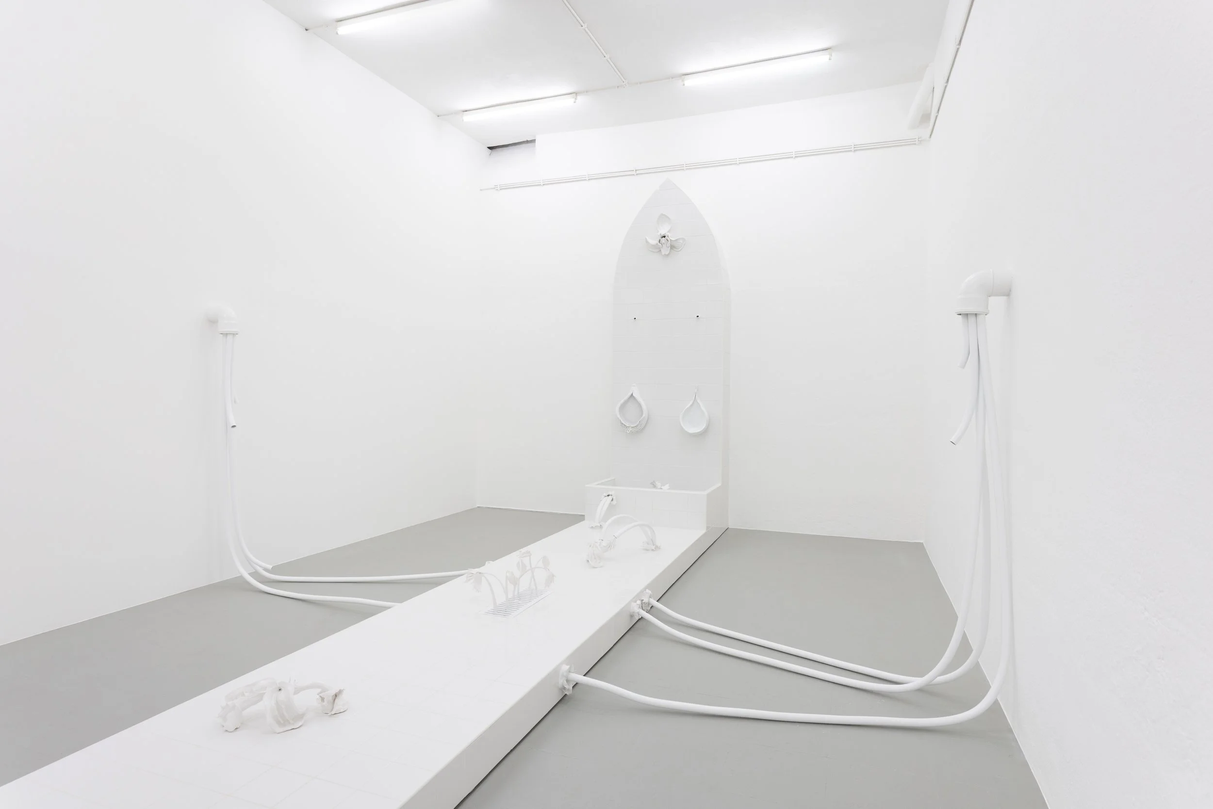 Art installation with white walls and floor, featuring white pipes, hoses, and objects arranged on a platform