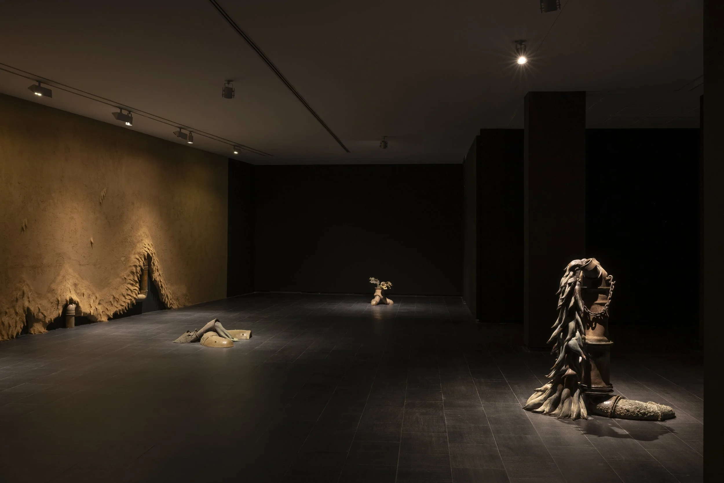 Empty art gallery with sculptures on the dark floor, a textured wall on the left, and black walls on the right. Overhead lights illuminate the sculptures, which include a broken column draped with leaves, a small figure with a flower, and a set of ob