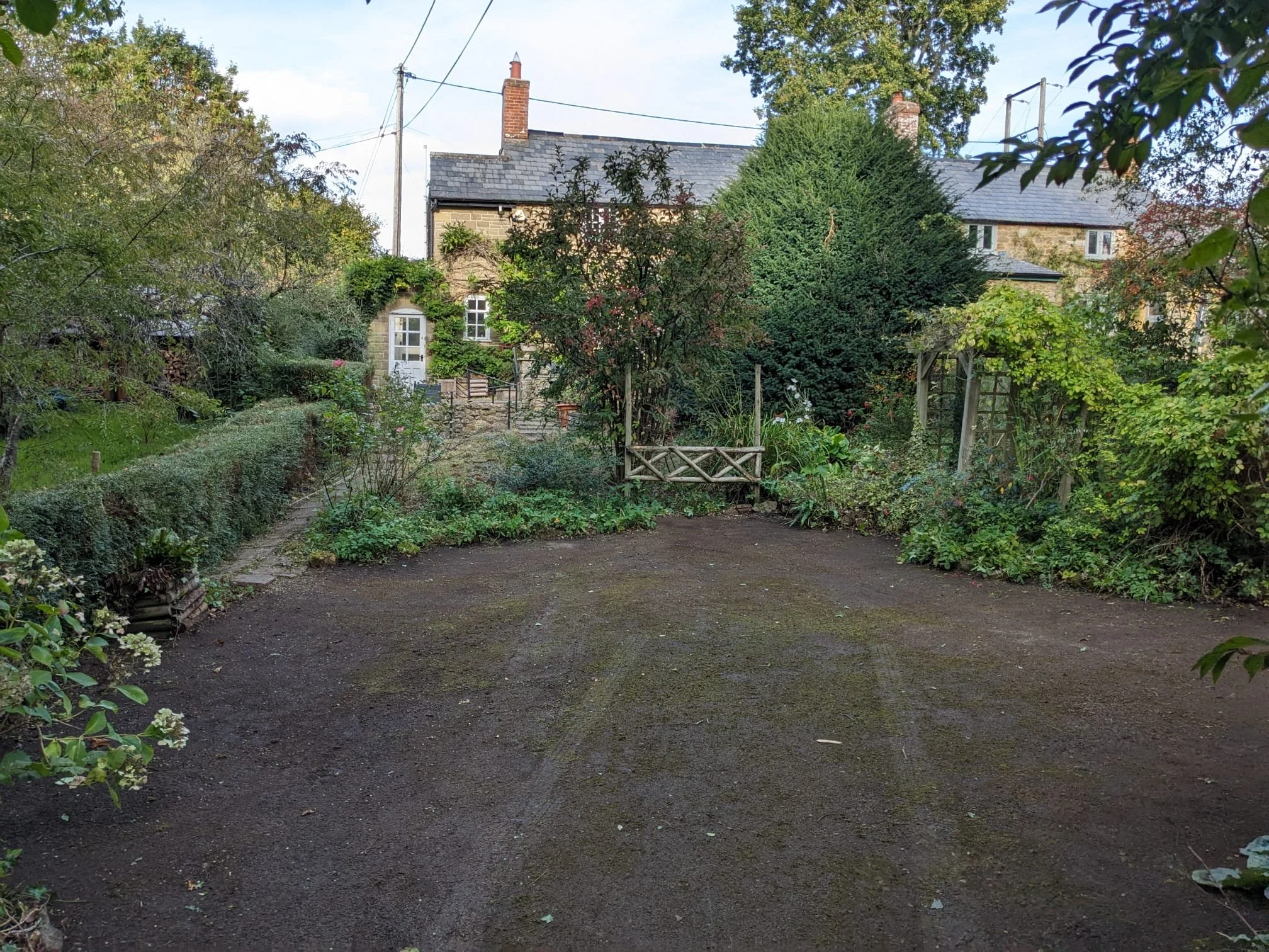 A gravel yard in front of a stone house with greenery and trees surrounding it, including a large bush and vine-covered door.