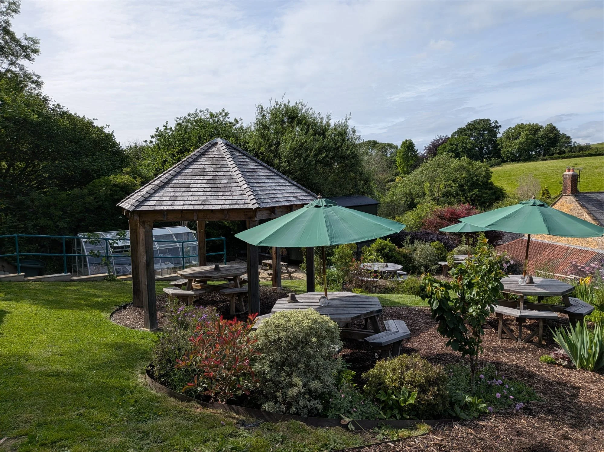 View of a backyard with green lawn, wooden gazebo, green umbrellas, picnic tables, surrounding trees, shrubs, and a hill with houses in the background under a partly cloudy sky.
