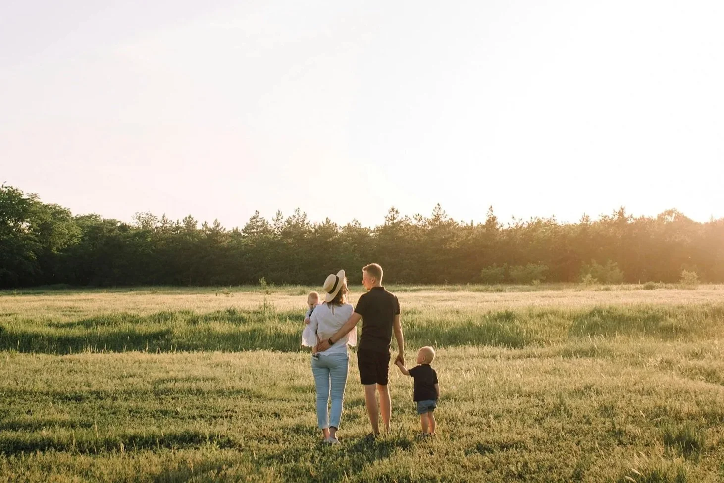 A family of four holding hands and walking in a grassy field during sunset, with trees in the background.