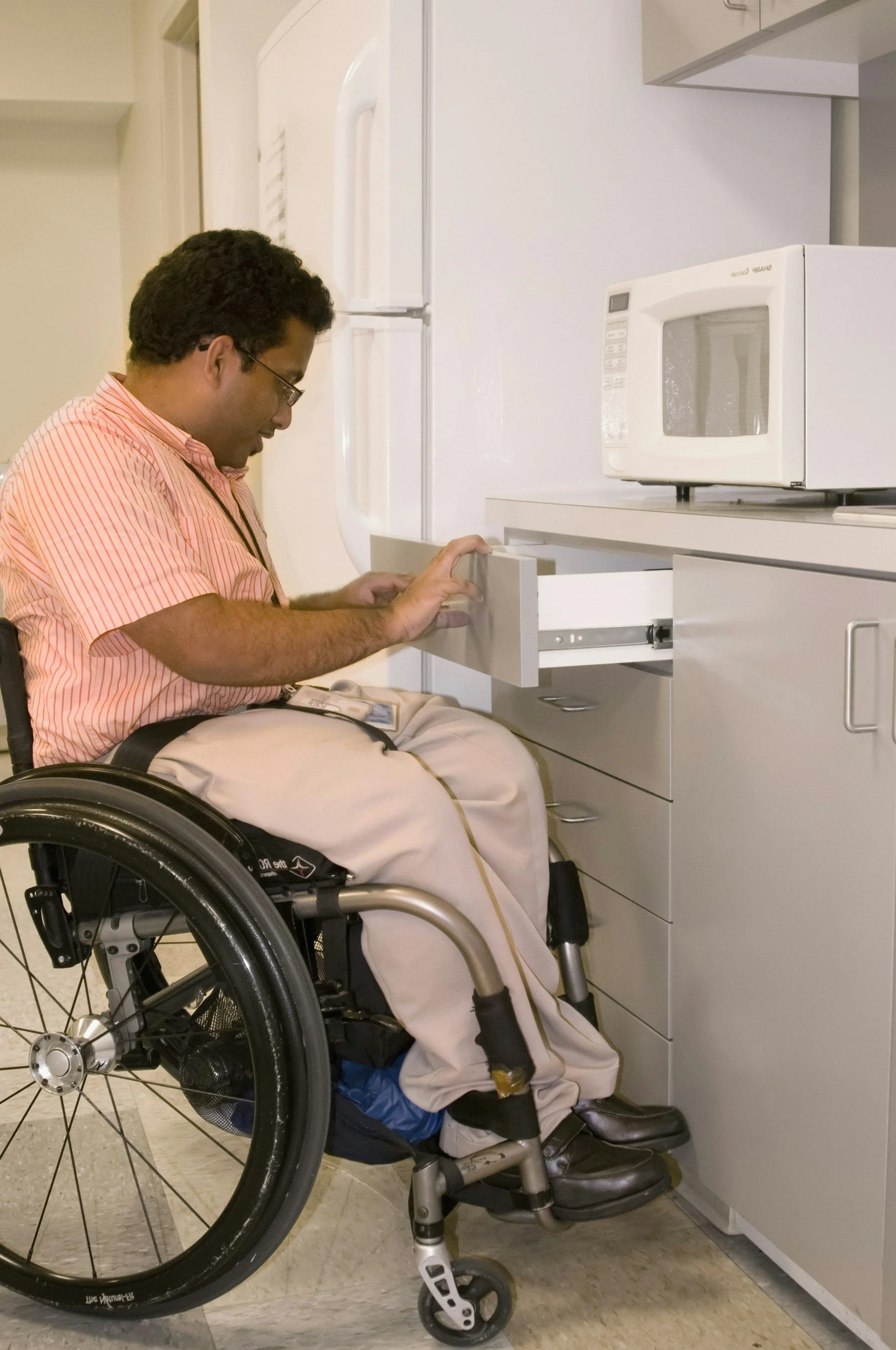 A man in a wheelchair opening a drawer in a kitchen. The man has dark curly hair and is wearing glasses, a tan and orange striped shirt, tan pants, and black shoes. The kitchen has white cabinets, a microwave on the countertop, and a white refrigerator.