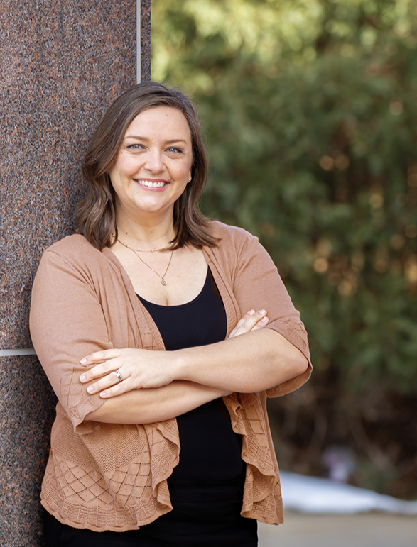 A woman with brown hair smiling, standing outdoors with her arms crossed against a brown brick wall and green trees in the background.