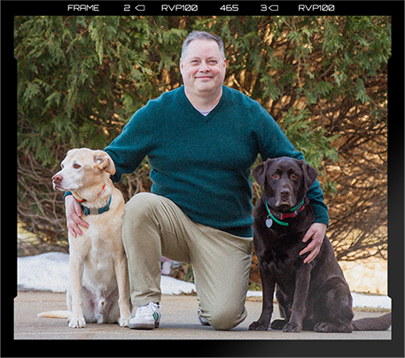 A man kneeling outdoors between two dogs, one light-colored and one dark-colored, with trees in the background.