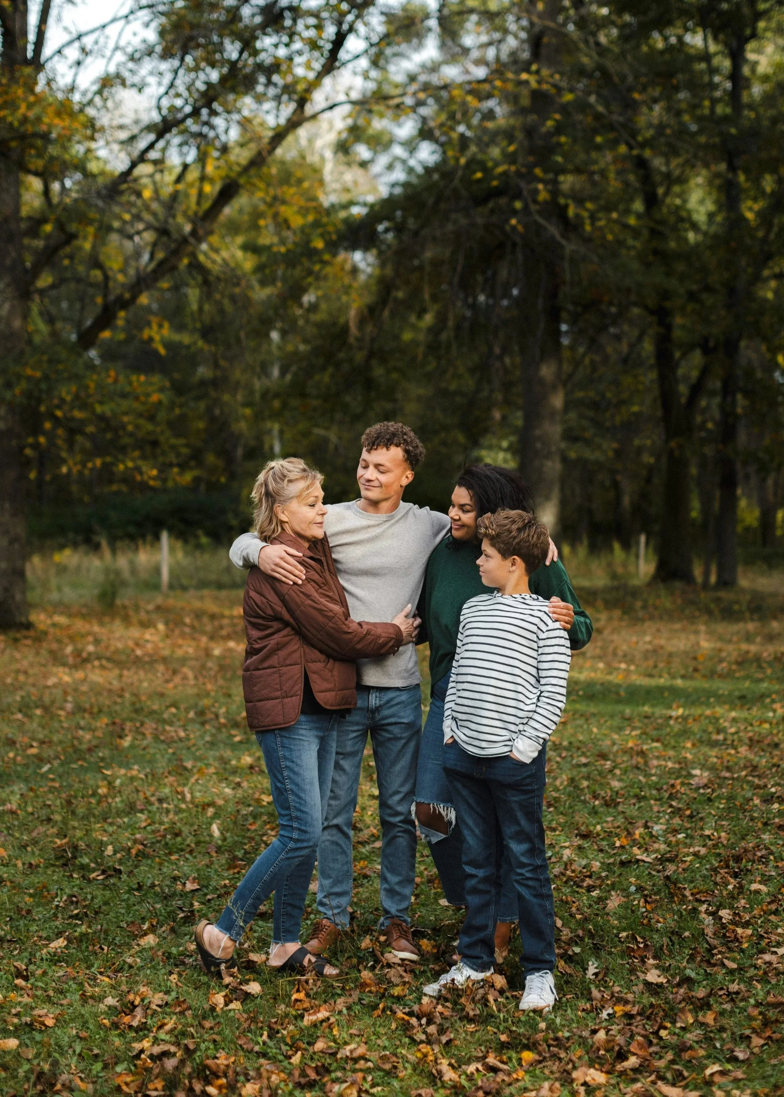 A group of five people, including children and adults, standing close together outdoors in a park surrounded by trees with autumn leaves, showing affection and warmth.