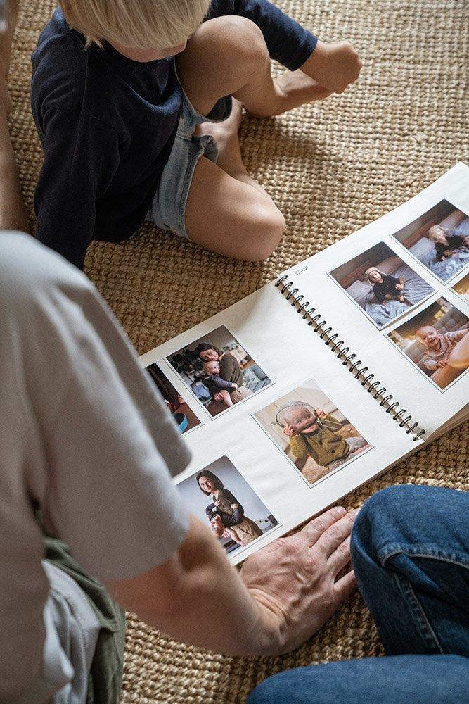 A young child sitting on a woven rug looking at a photo album with an adult, who is pointing at photos of children in the album.
