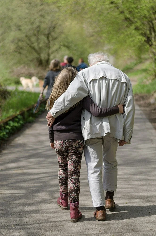 An elderly man and a young girl -- grandpa and granddaughter -- walk arm in arm on a park path surrounded by trees, with a dog and other people in the background.